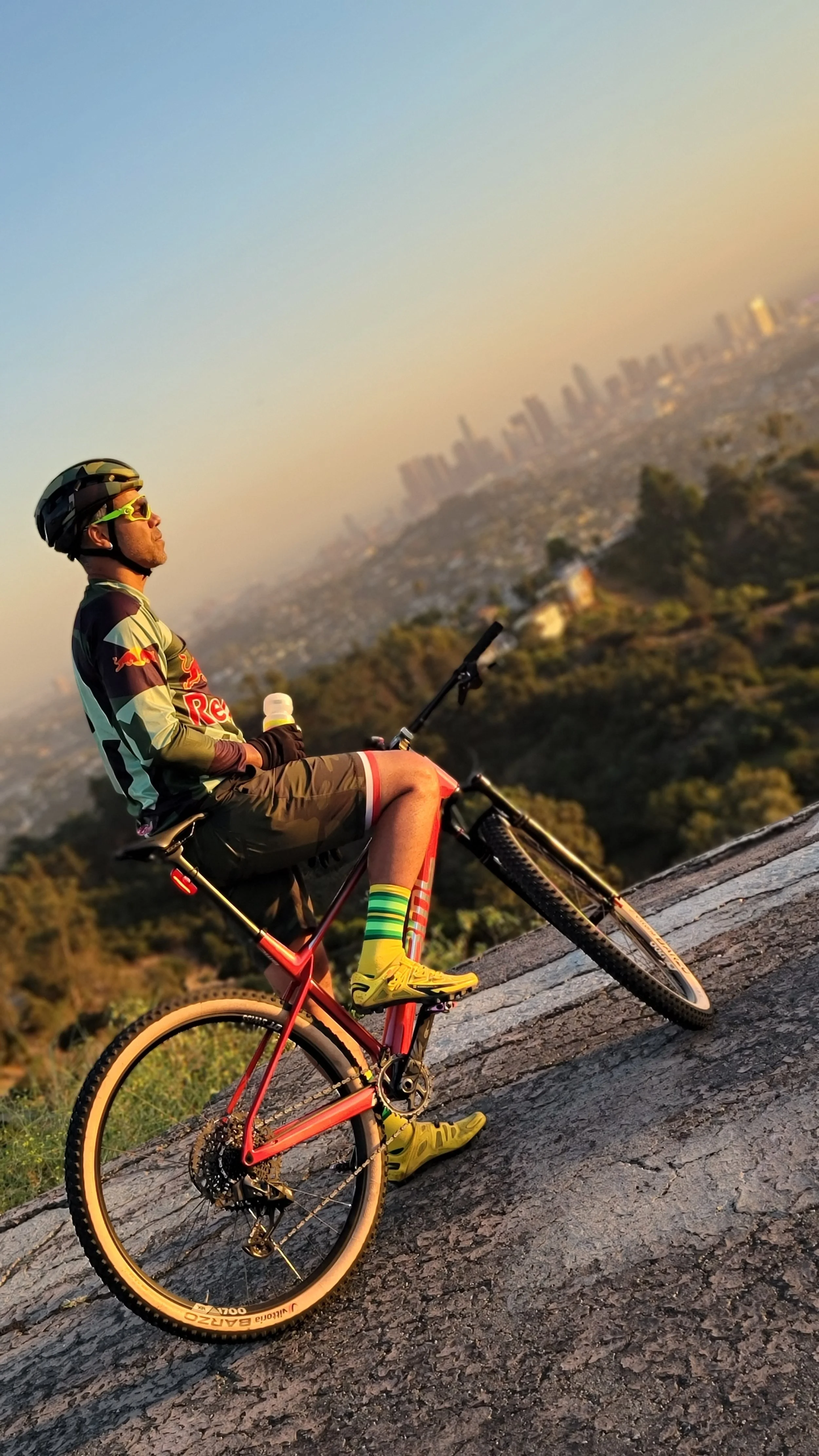 A man wearing a helmet, sunglasses, and a Red Bull cycling jersey sitting on his mountain bike on a hillside at sunset with a city skyline in the background.