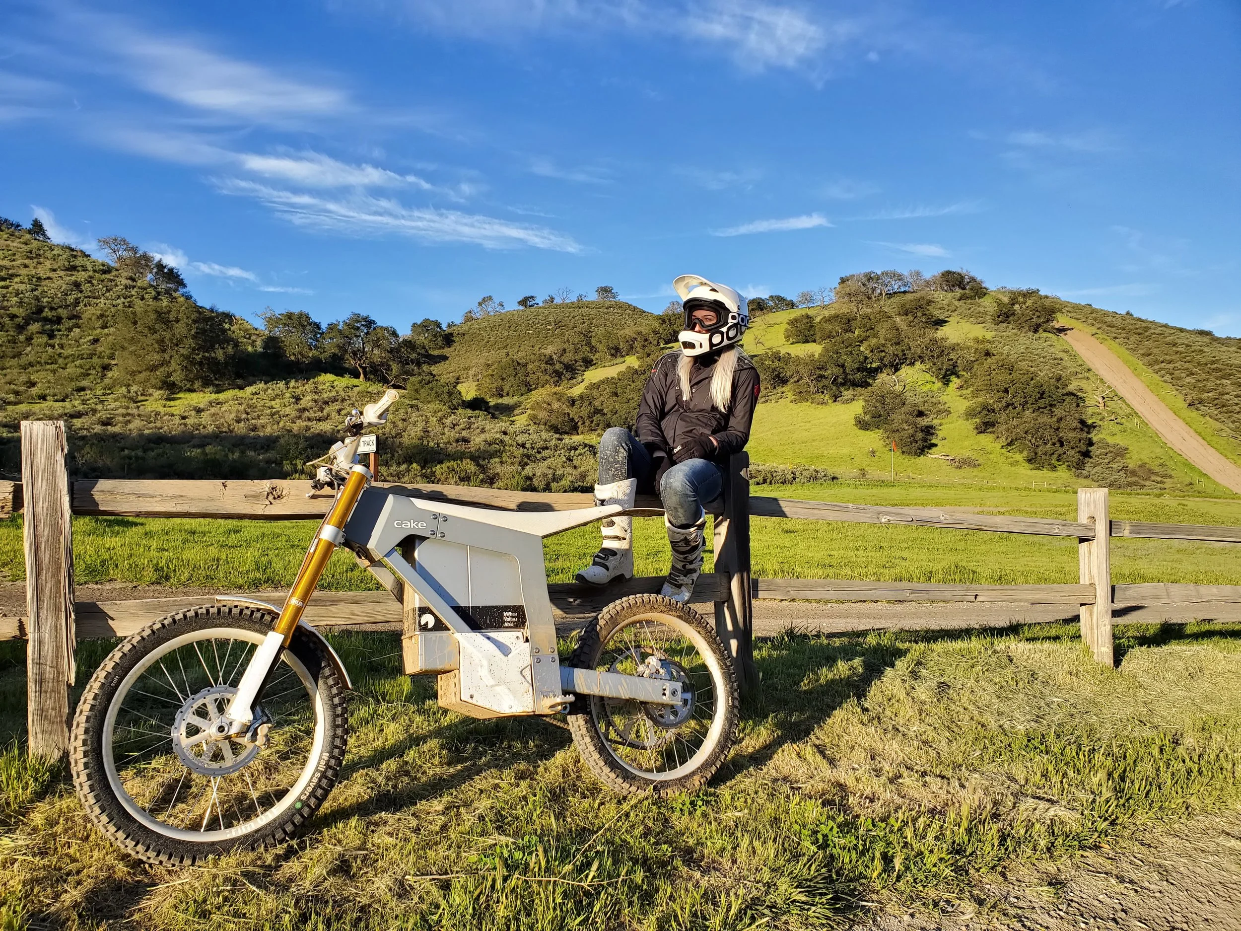 A woman sitting on a wooden fence with a mountain bike, wearing a helmet and riding gear, in a grassy field with green rolling hills and a blue sky in the background.
