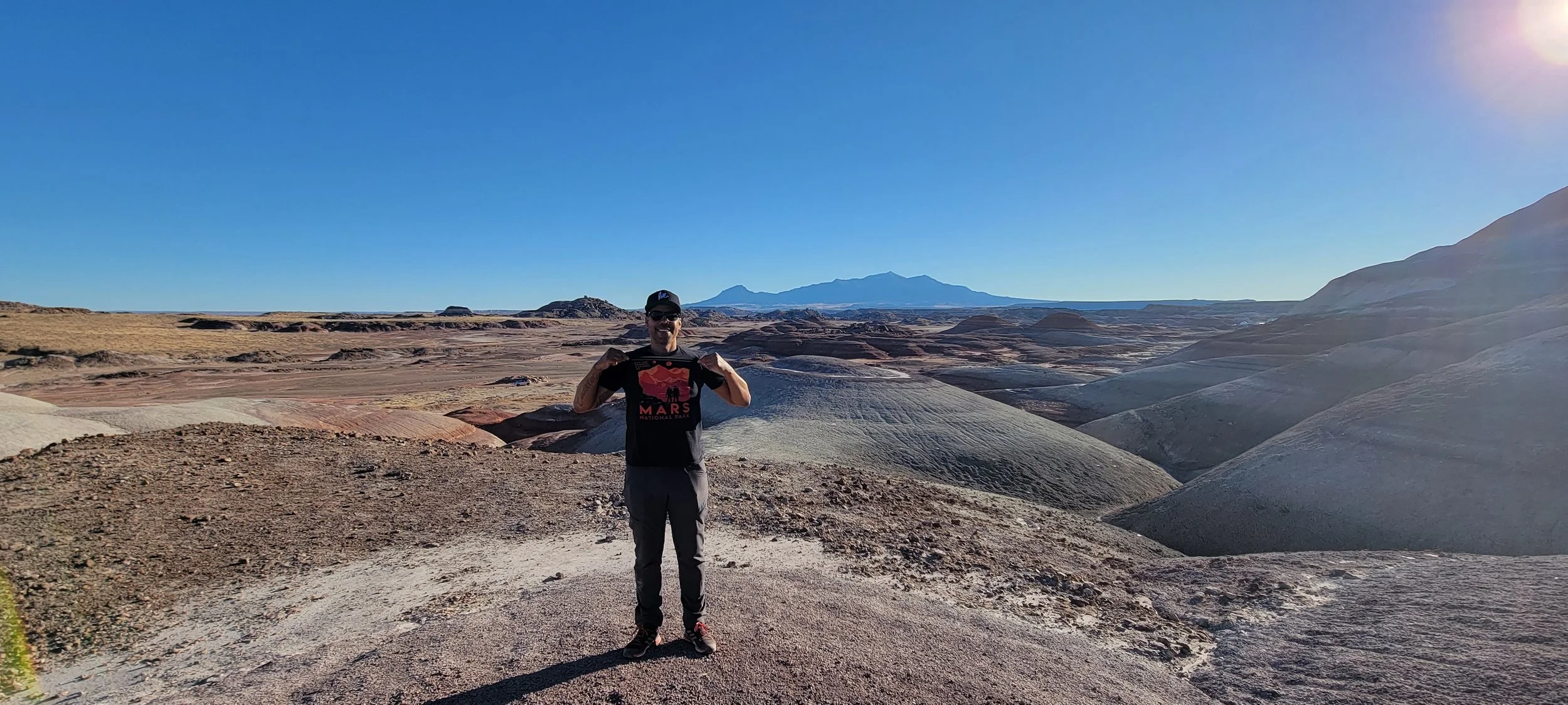 A person standing outdoors in a rocky desert landscape, holding a sign that says 'Mars' with mountains and a blue sky in the background.