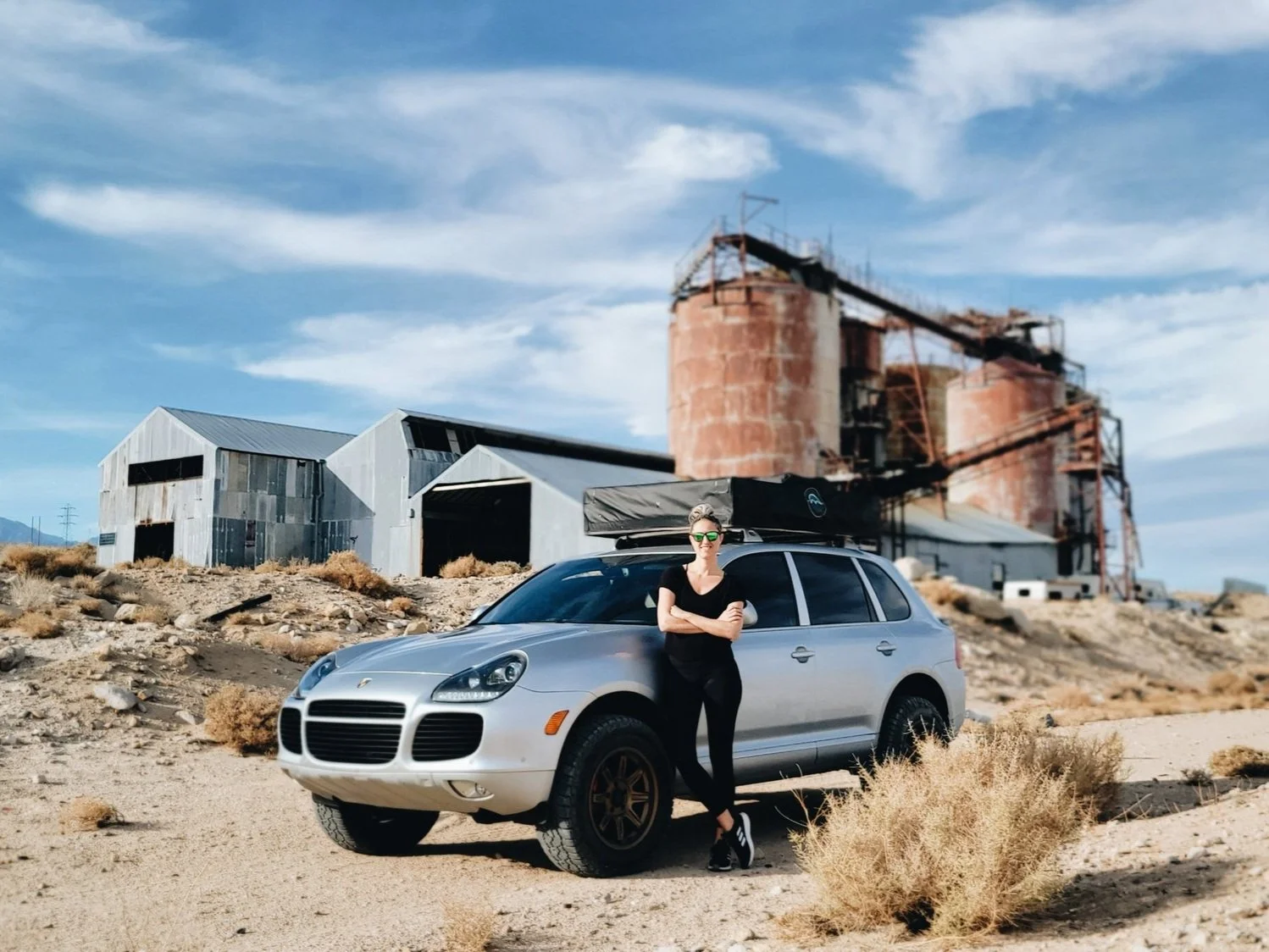 A woman with blonde hair, sunglasses, black outfit, standing with crossed arms, leaning against a silver Porsche SUV in a desert landscape with abandoned industrial buildings and rusty silos in the background.