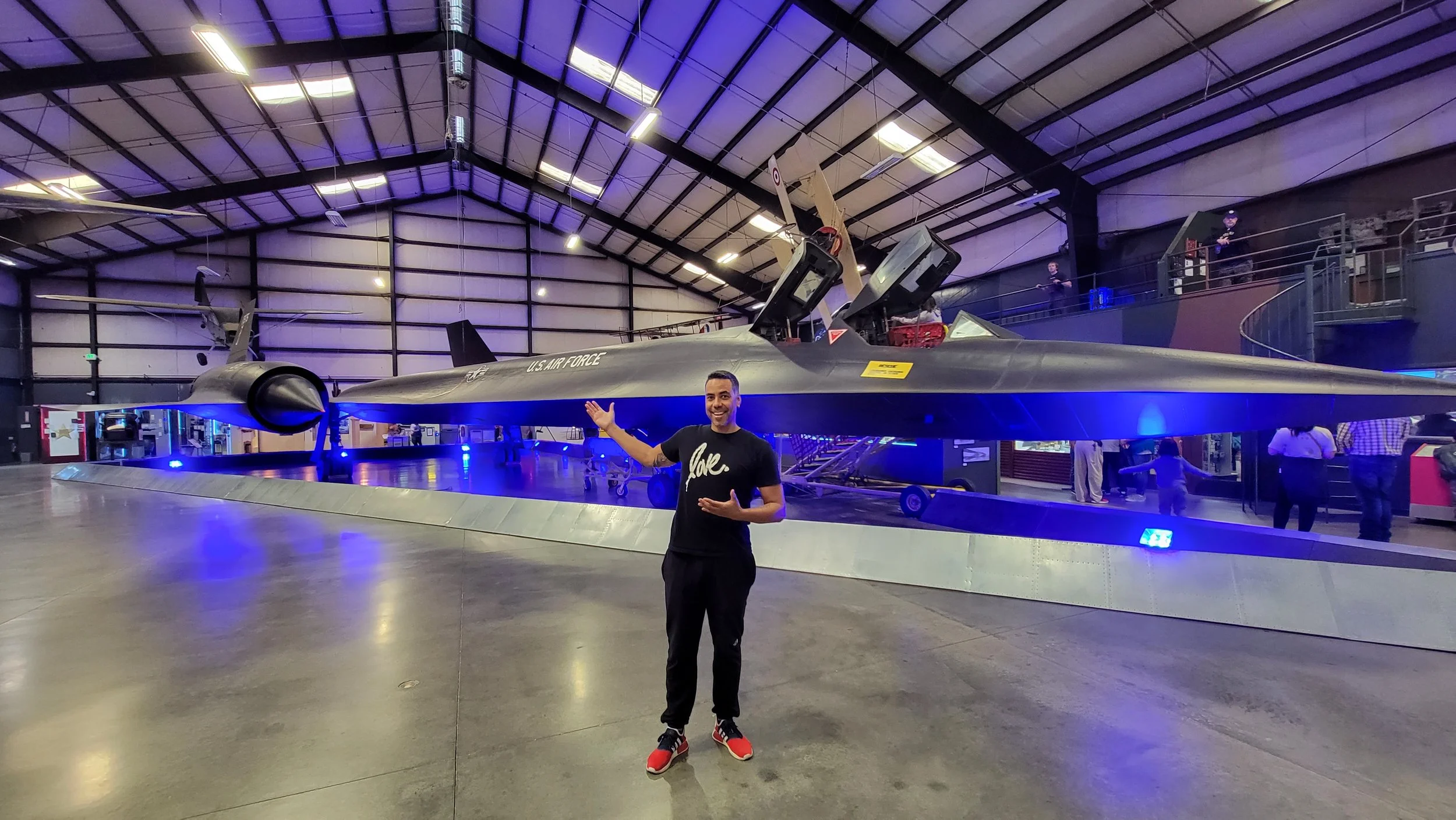 A man standing in front of a stealth fighter jet displayed at an indoor museum, with people in the background.