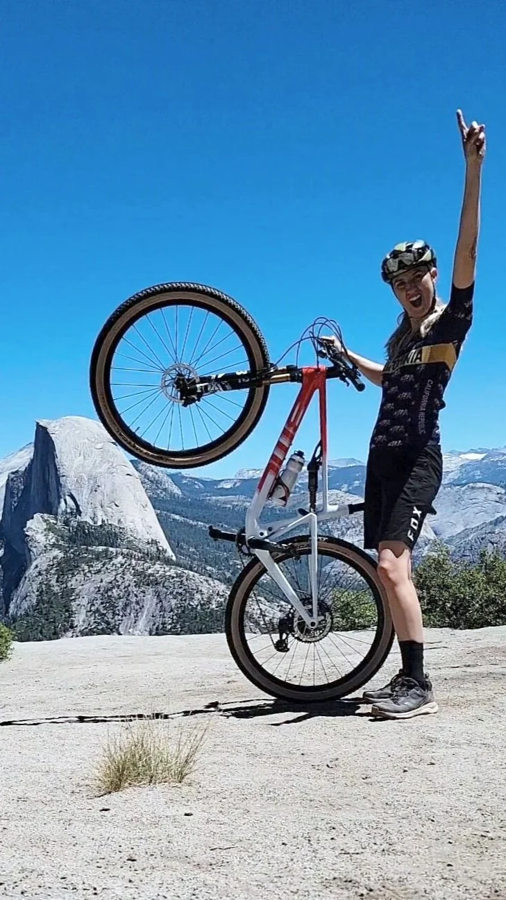 A woman in outdoor gear and a helmet holding a mountain bike upright on a sandy surface with mountainous landscape in the background under a clear blue sky.