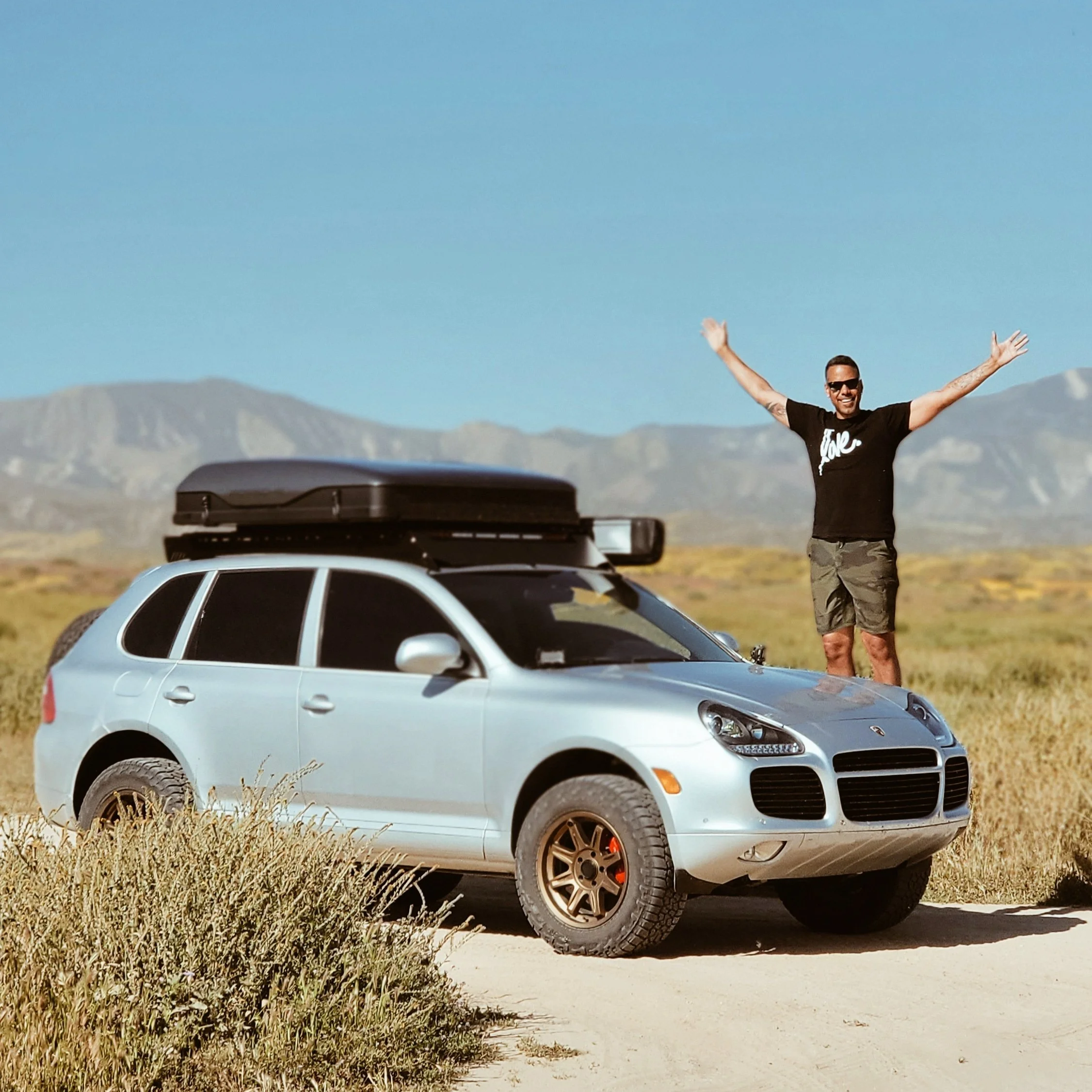 A man standing on a dirt road in a desert landscape with mountains in the background, smiling and raising his arms next to a silver SUV with a black roof box.