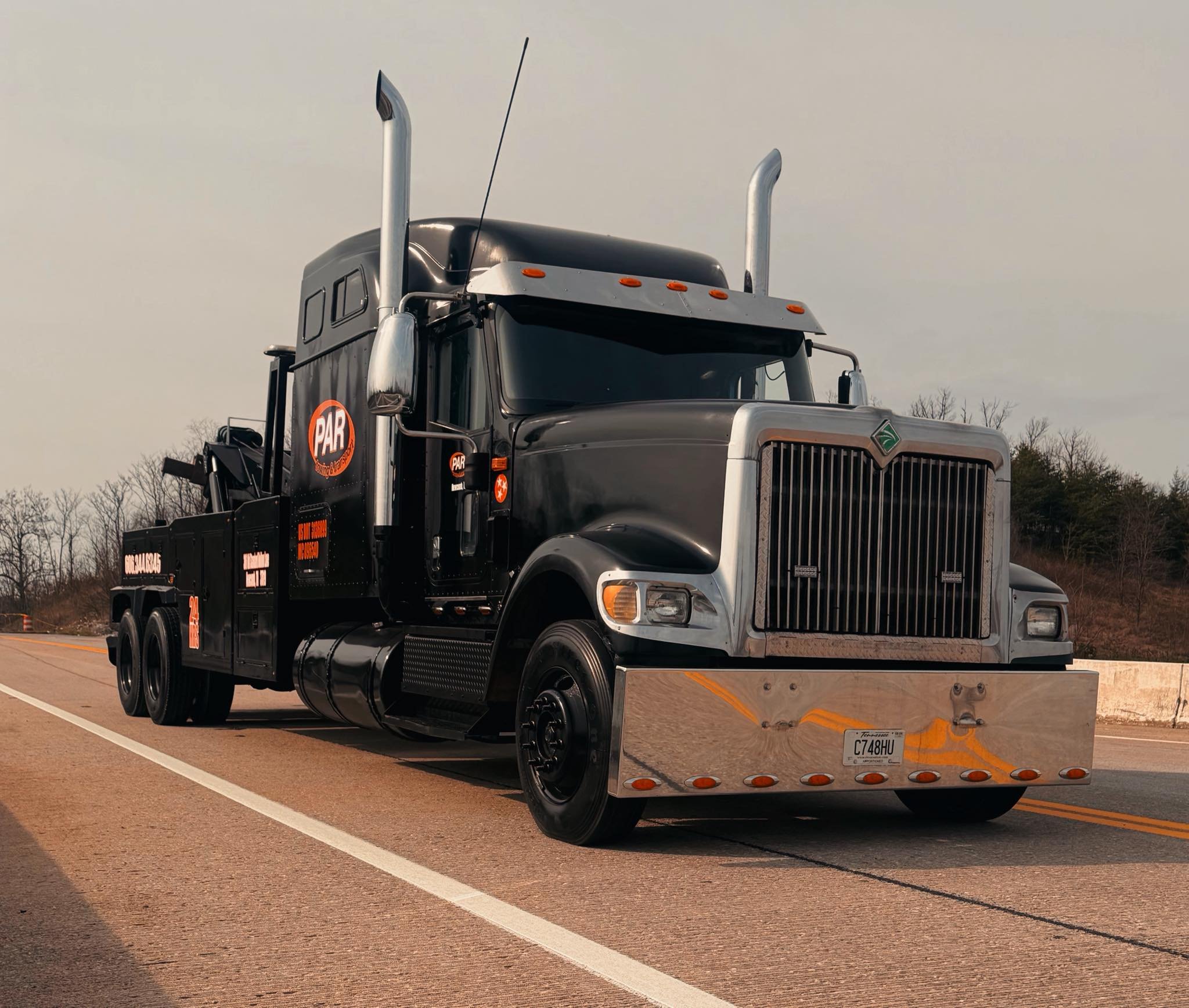 A black semi-truck with a silver front grill parked on the side of a road. The truck has an orange and white logo on the door and a Tennessee license plate. The truck has tall vertical exhaust pipes and a silver sun visor over the windshield. The background shows leafless trees and a partly cloudy sky.