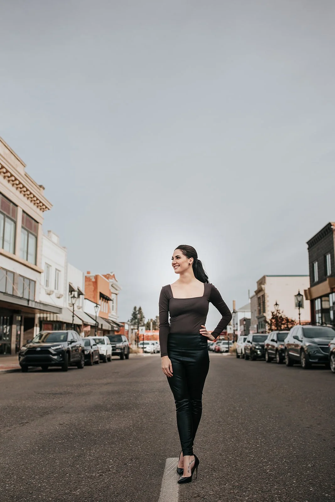 Mackenzie Johnson real estate agent in black outfit and high heels standing in the middle of a downtown Medicine Hat, smiling, with buildings and cars around.