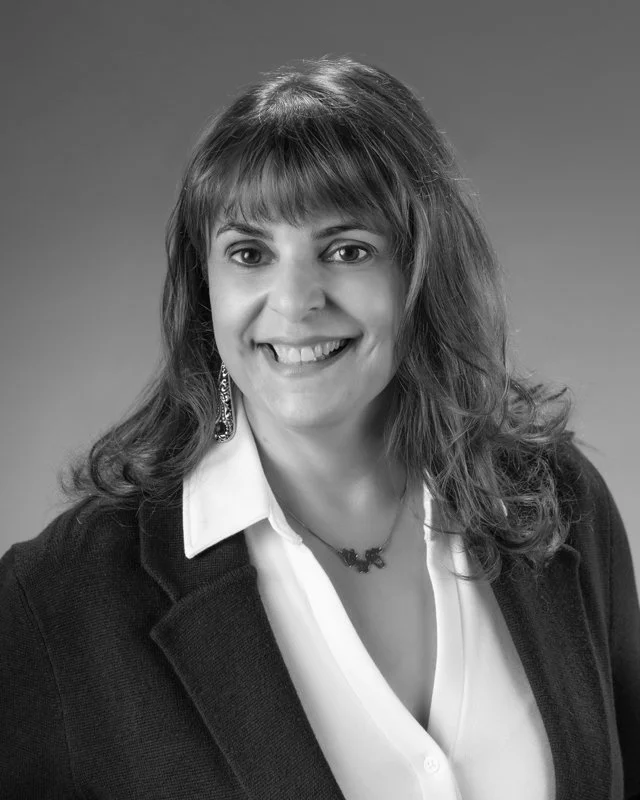 Black and white portrait of a woman with shoulder-length wavy hair, smiling, wearing a white shirt, dark blazer, and earrings.