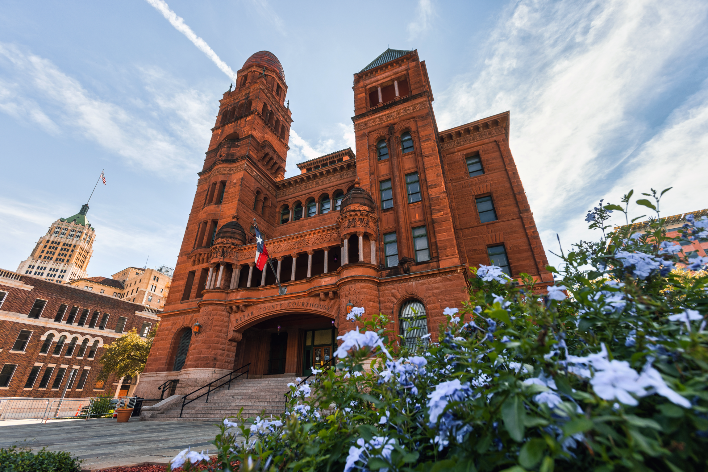 Red brick courthouse with a tall clock tower, flags, and steps leading up to the entrance, surrounded by greenery and other buildings under a partly cloudy sky.