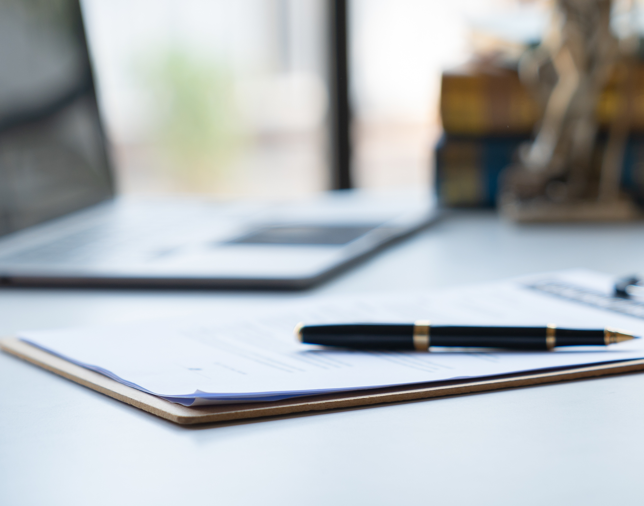 Close-up of a black and gold pen on top of a clipboard with papers, on a white desk, with blurred office background.