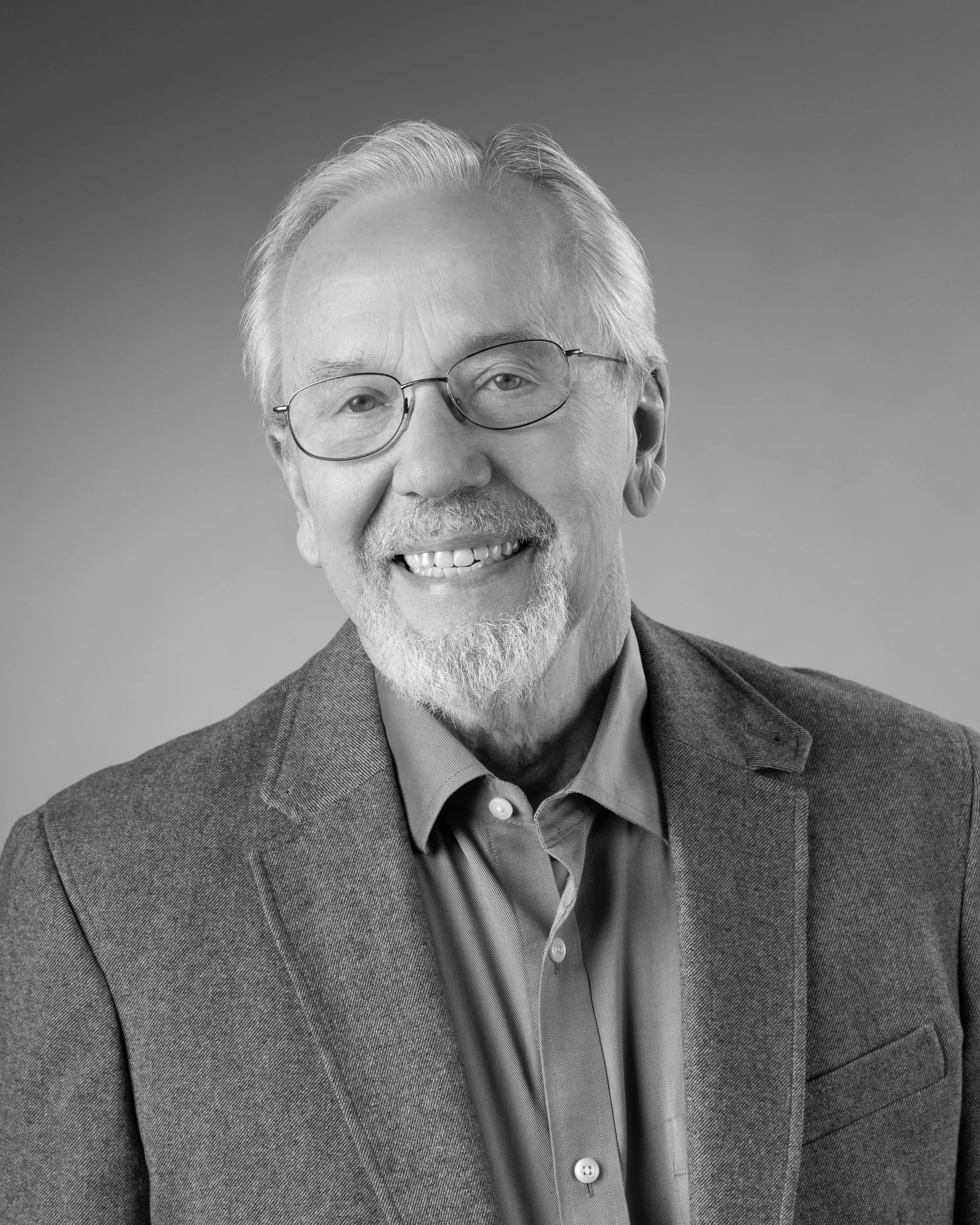 Black-and-white portrait of a smiling older man with glasses, gray hair, and a beard, wearing a suit jacket and a collared shirt.