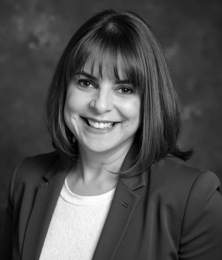Professional headshot of a smiling woman with shoulder-length hair, wearing a dark blazer and light top against a blurred background.