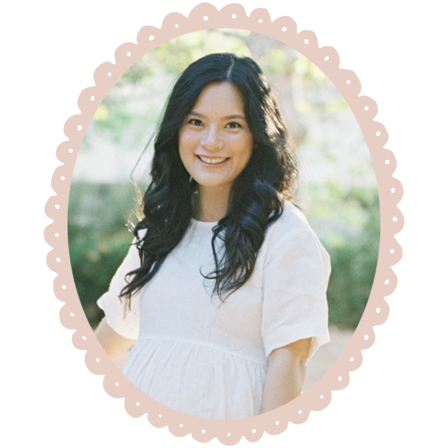 A smiling woman with long, wavy black hair wearing a white dress, standing outdoors with a blurred background of trees and greenery.