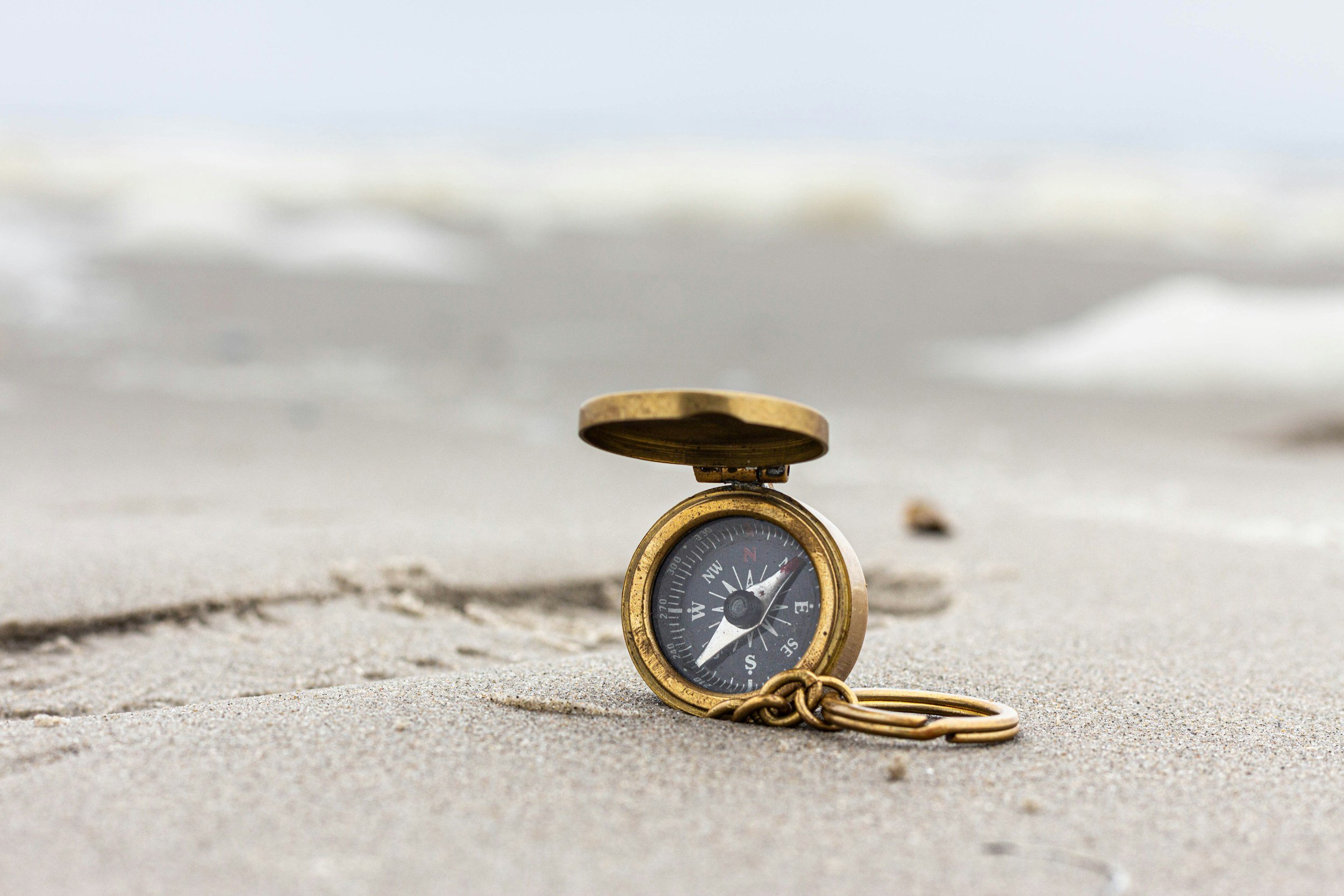 A vintage brass compass with a chain, resting on sandy beach near the shoreline.