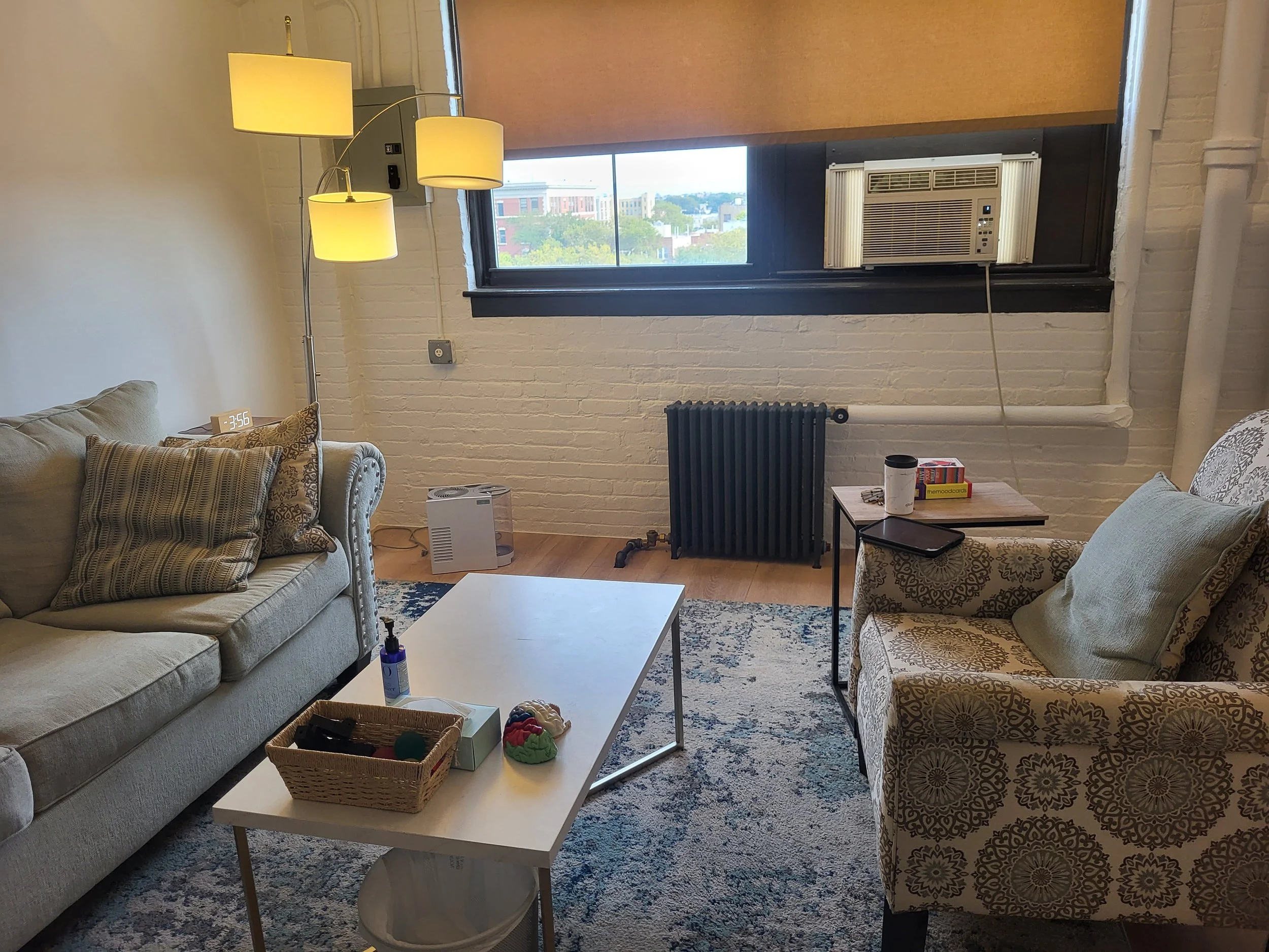 Living room with beige sofa on the left, patterned armchair on the right, white coffee table in the center, blue and white area rug, window with an air conditioning unit, radiator, floor lamp, and various items on tables and walls.