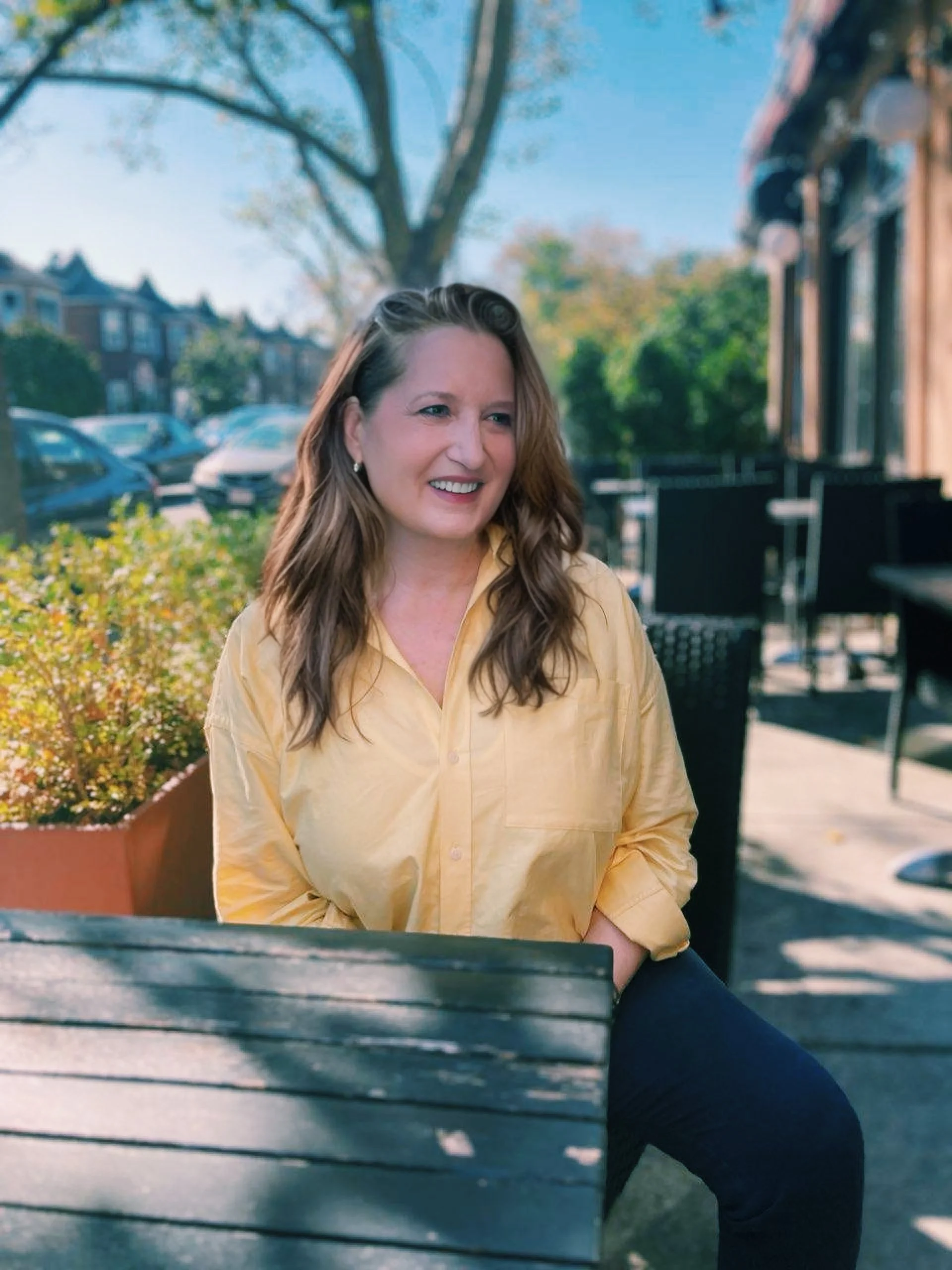 A woman with long brown hair smiling, wearing a yellow shirt, sitting outdoors on a patio on a sunny day with trees and parked cars in the background.