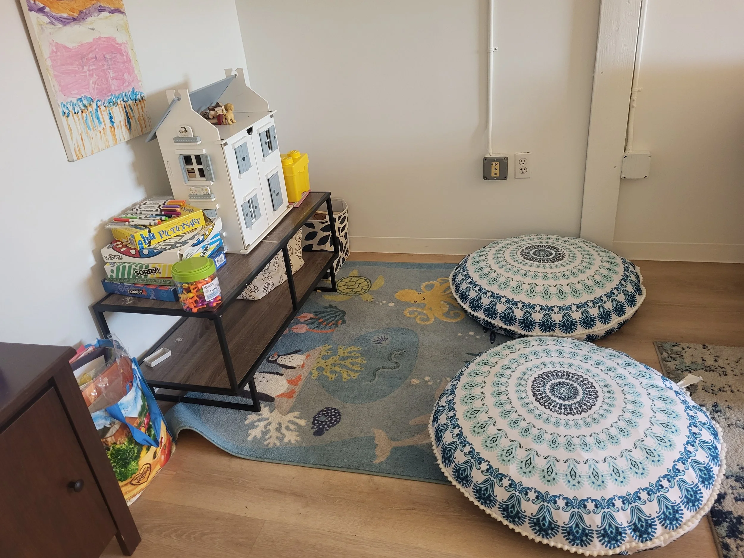 Children's playroom with a colorful rug, two large round cushions with intricate blue and white patterns, a black metal shelf with toys, board games, and books, and a white dollhouse on top.
