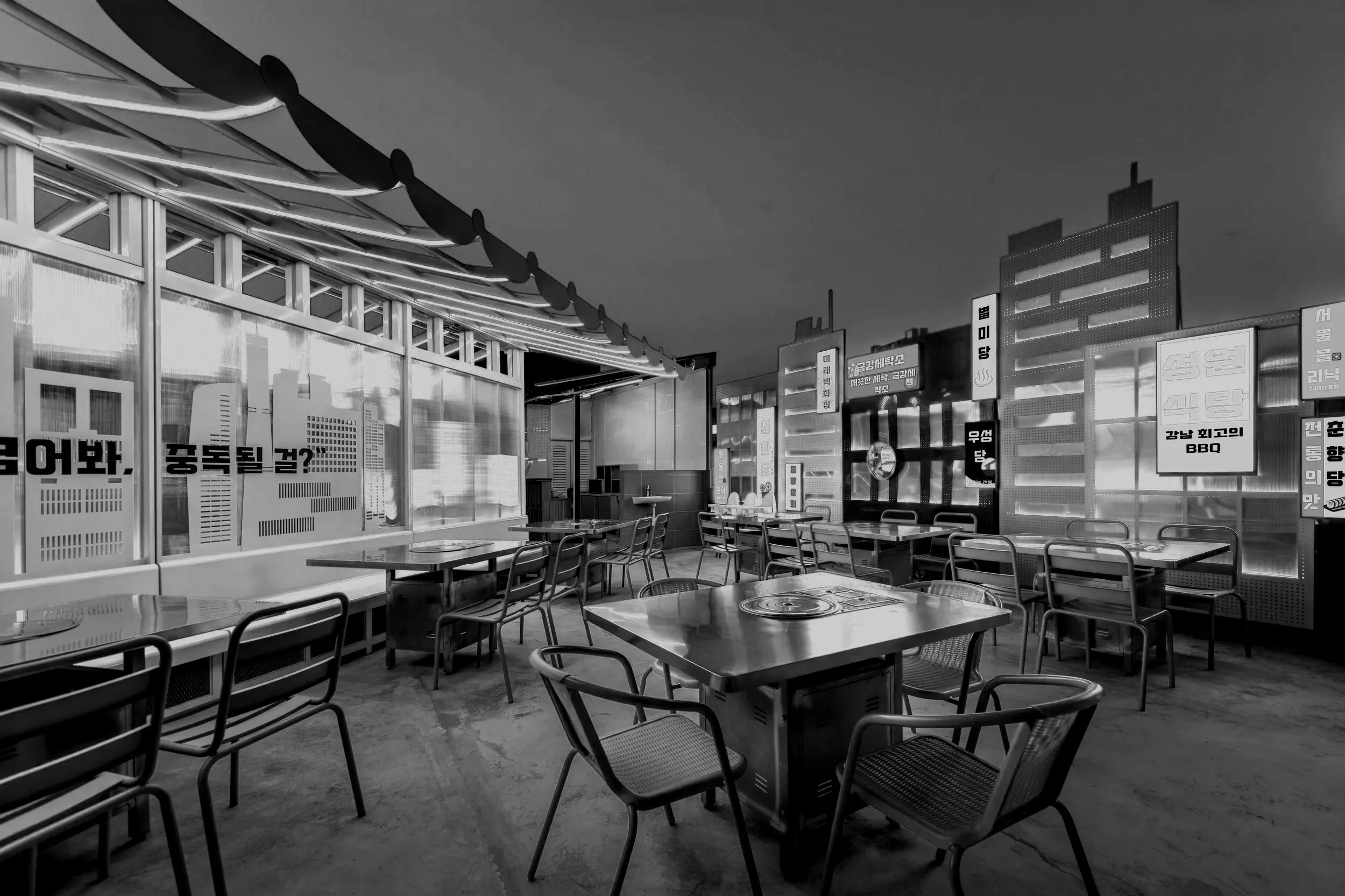 jibungae kbbq. Rooftop restaurant or patio with tables and chairs, city skyline in the background, illuminated signs in Korean, and decorative elements along the edge of the roof.