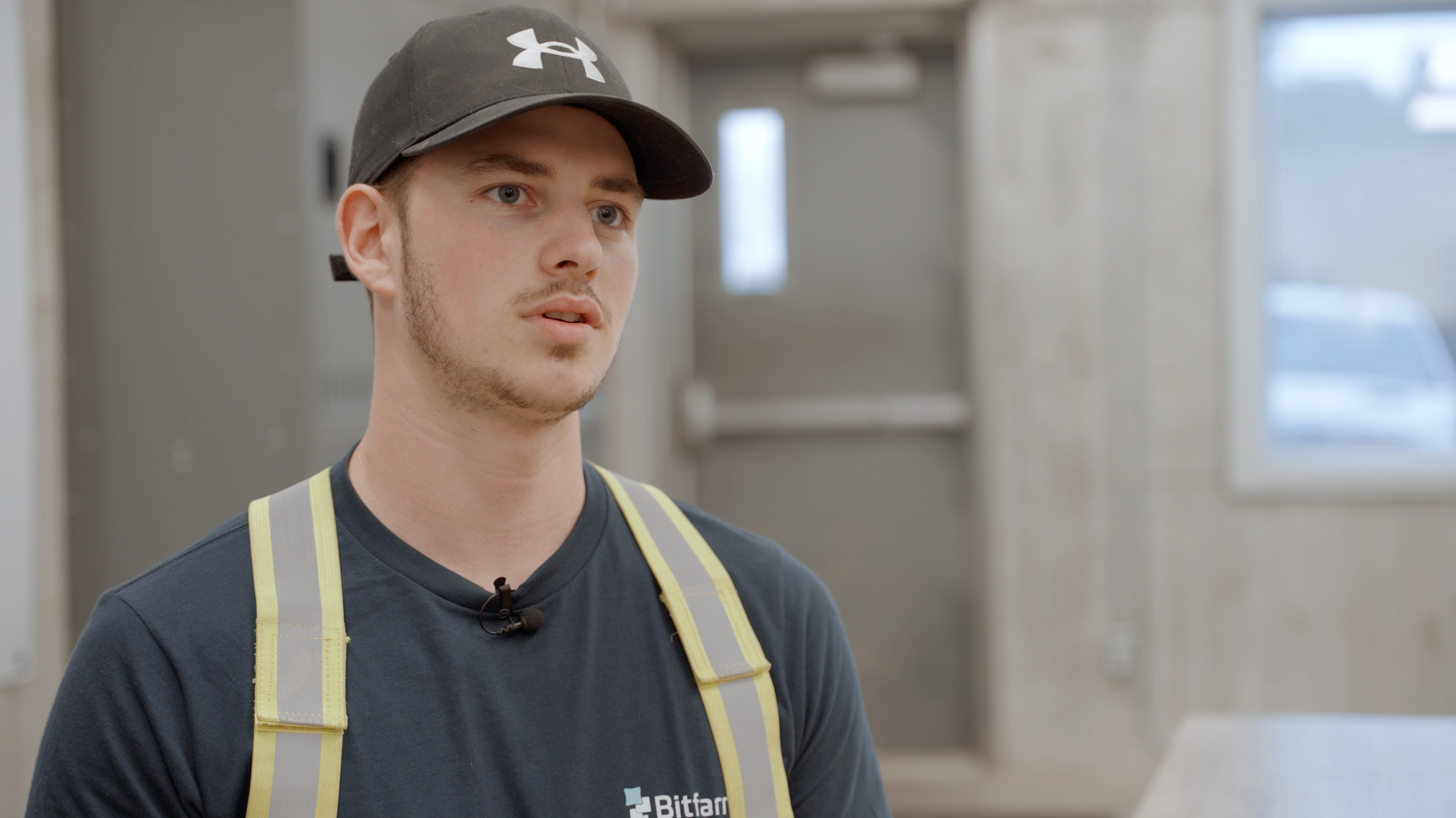 A young man wearing a black Under Armour cap, a dark t-shirt, and a high-visibility safety vest, standing indoors with a serious expression.