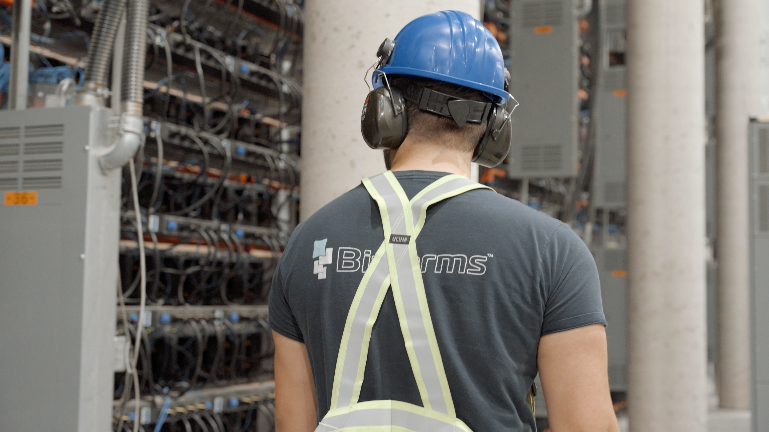 Back view of a worker wearing a blue safety helmet, yellow safety harness, and black t-shirt in a server room with racks of electronic equipment and wiring.