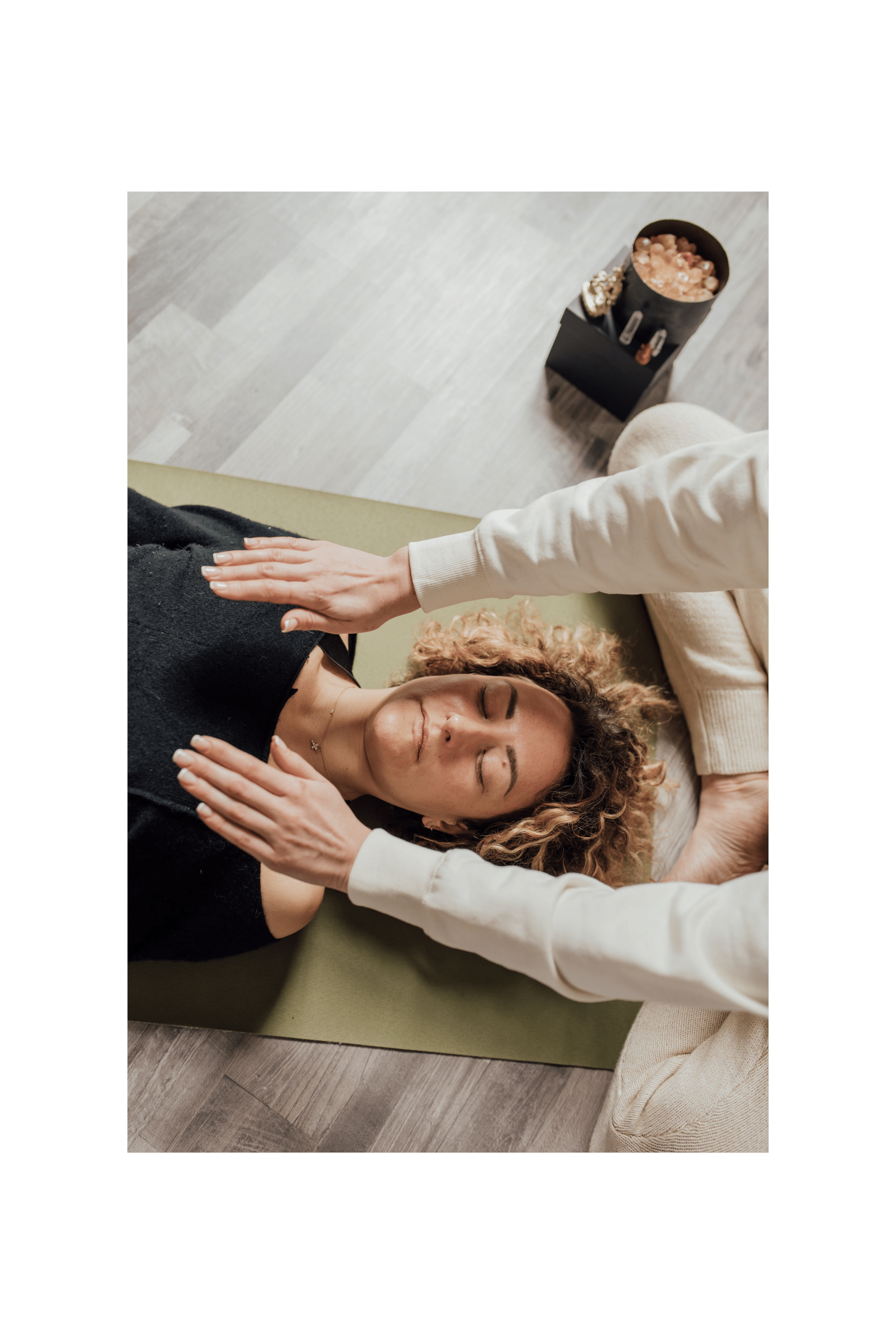 Une femme à cheveux bouclés étendue sur un tapis vert, recevant un massage, avec une personne en train de lui masser la poitrine. La scène se déroule dans une pièce avec un plancher en bois clair et une personne portant un pantalon beige et un haut blanc est visible à droite.