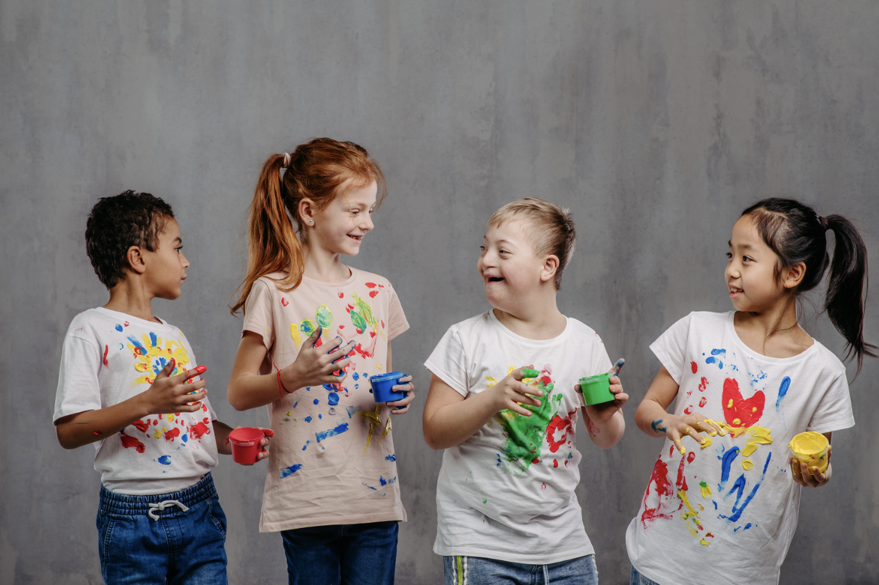 Children in paint-covered T-shirts hold paint pots and smile at each other, representing inclusive, creative school environments where every child is seen, supported and able to participate.
