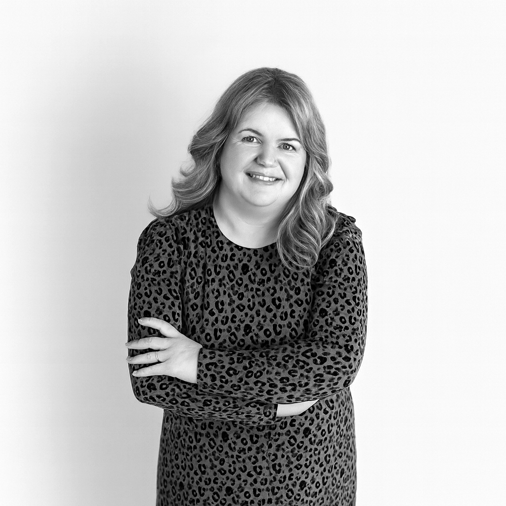 Black and white portrait of a woman with shoulder-length wavy hair, smiling, wearing a long-sleeve leopard print top, standing against a plain wall.
