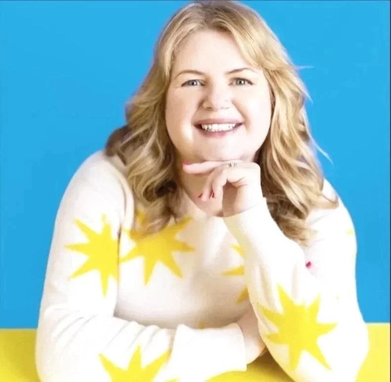 Young woman with blonde hair smiling at the camera, seated at a table with a yellow and blue background, wearing a white sweater with yellow star patterns.