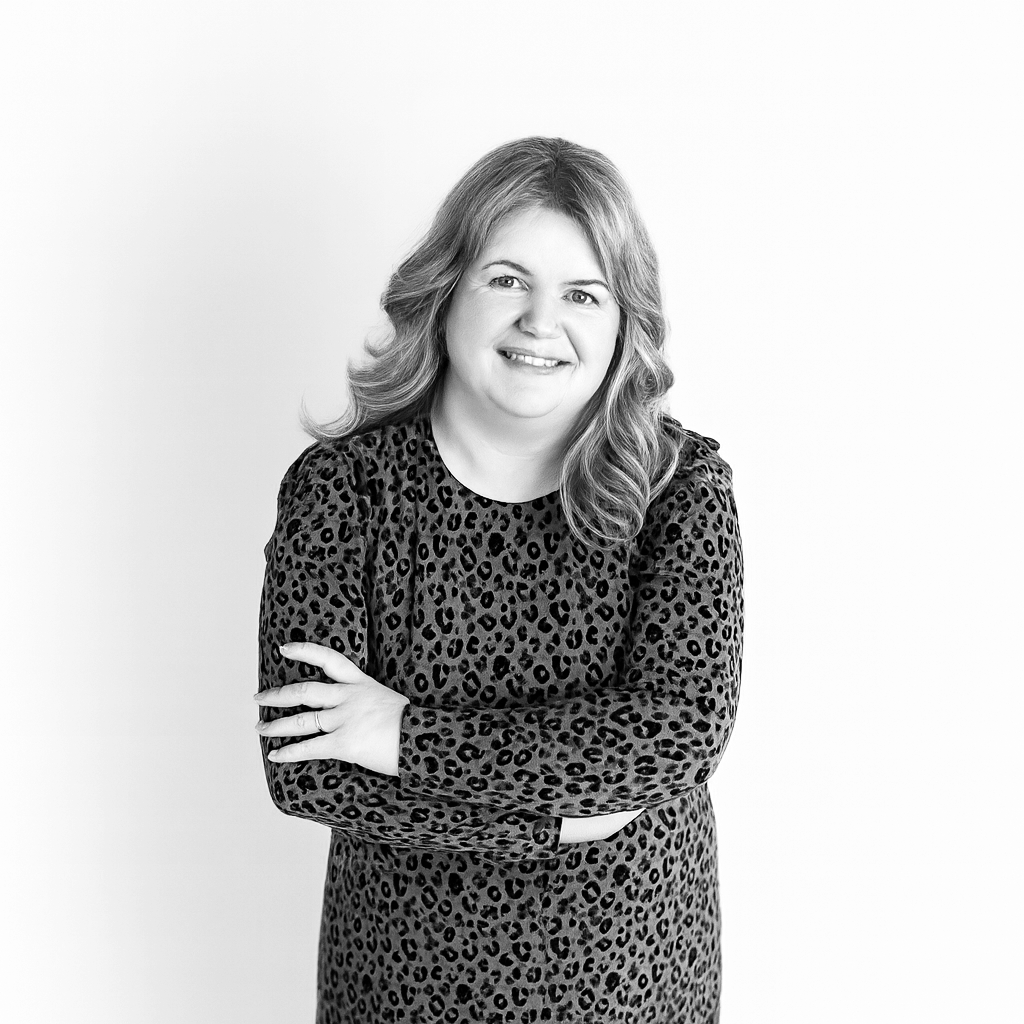 Black and white portrait of a smiling woman with wavy hair, wearing a leopard print long-sleeve top, with arms crossed, against a plain background.