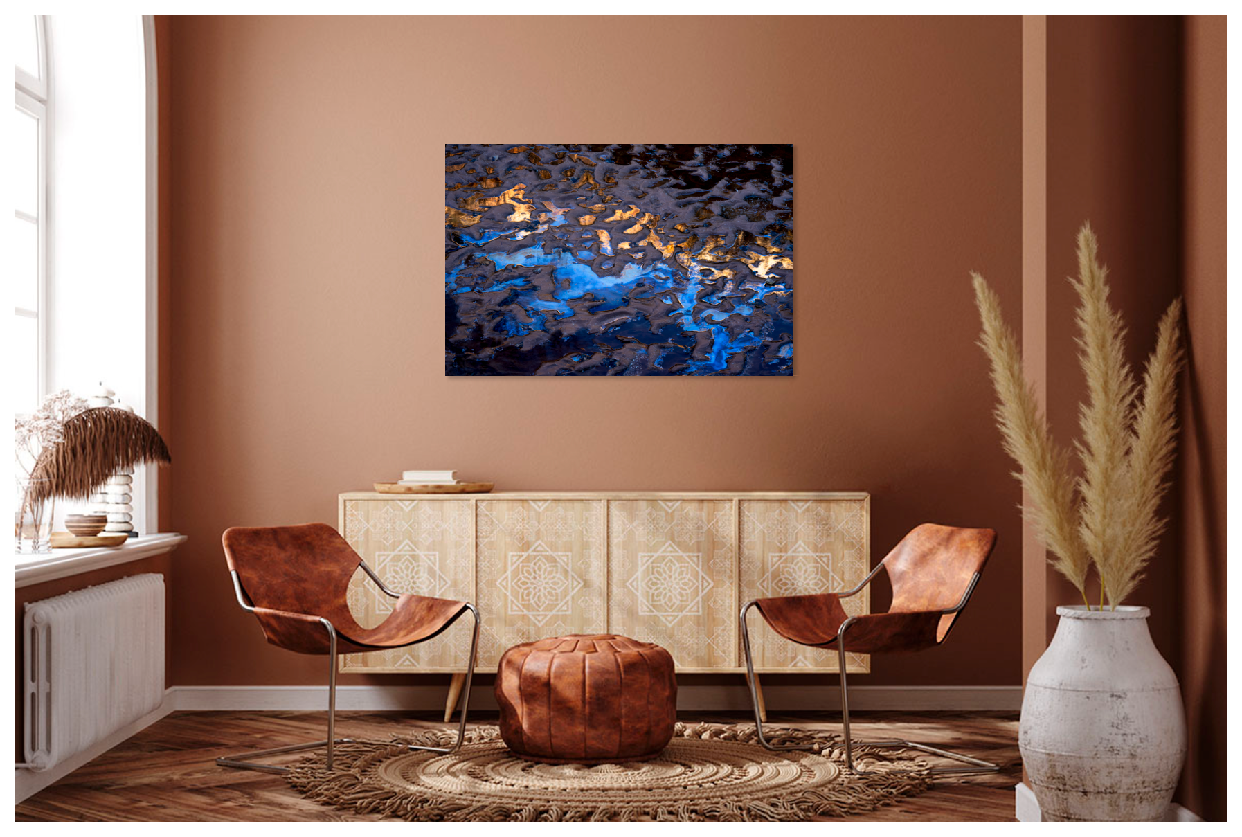 Interior of a cozy living room with a brown wall and natural light from a window. Two brown leather chairs face each other with a round leather ottoman between them. A decorative rug is underneath. A detailed cream-colored cabinet is behind the chairs. A large white vase with pampas grass stands in the corner. A modern art piece with blue, gold, and dark tones hangs on the wall.