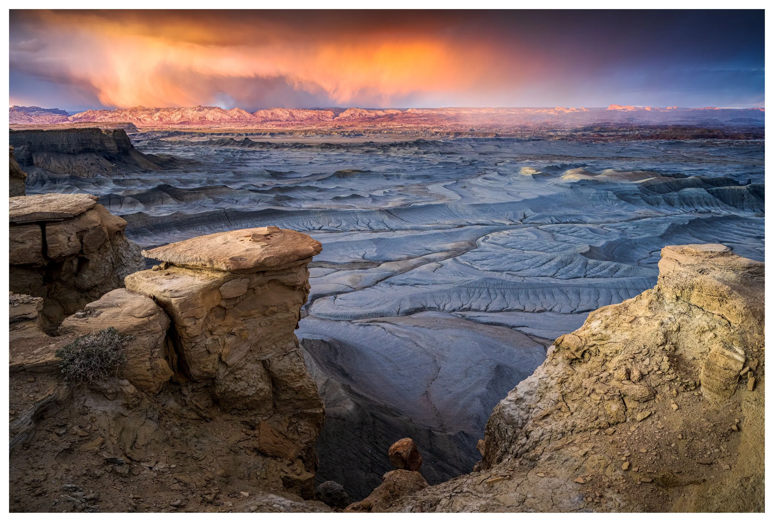 A vast desert landscape with eroded rock formations in the foreground and a colorful sunset sky with clouds in the background