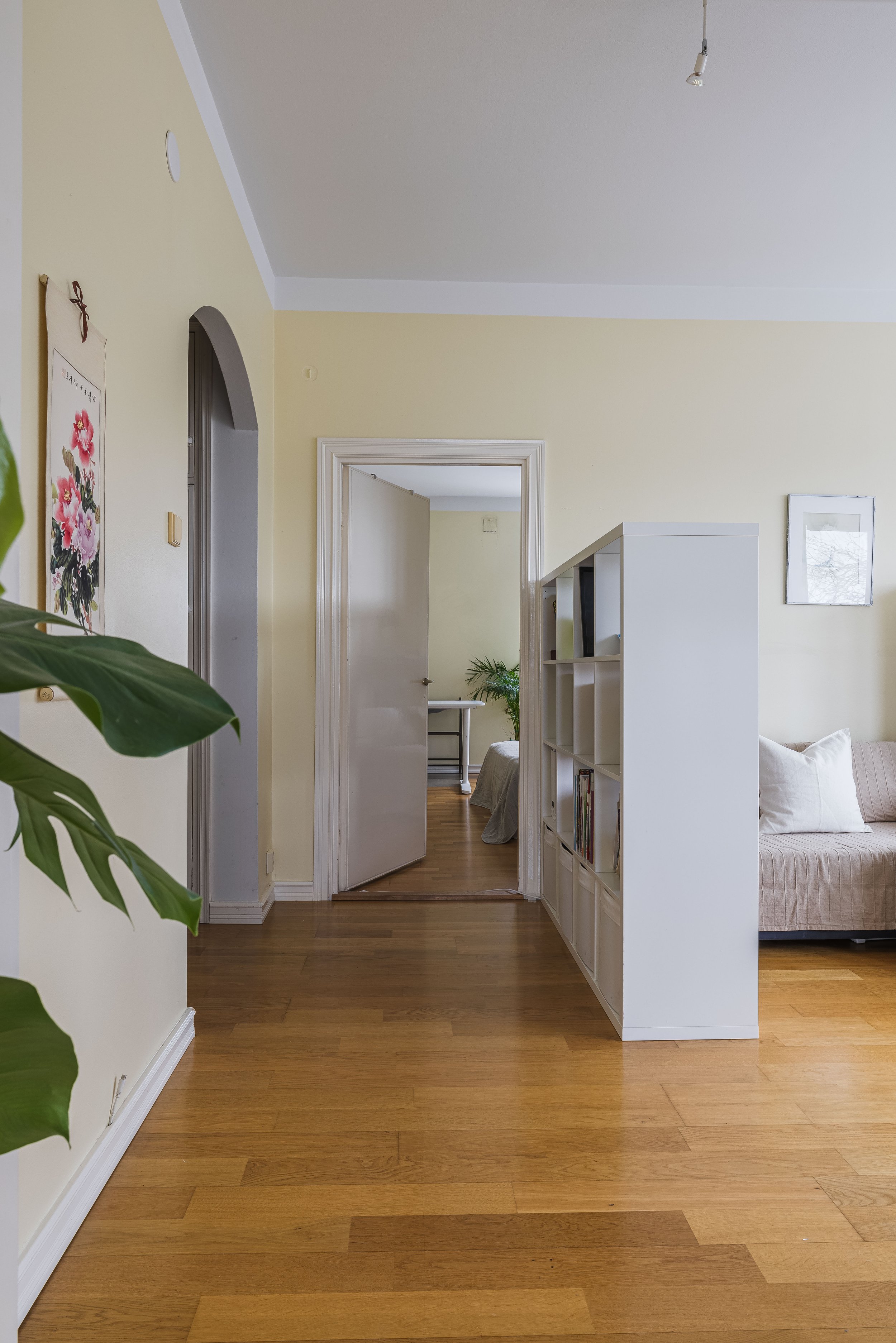 Modern living room with beige walls, wooden floor, white shelving, and a doorway leading to a bedroom.