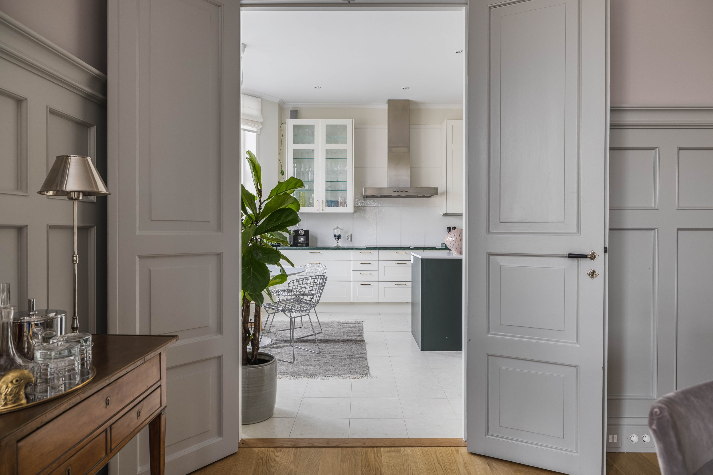View through an open door into a modern kitchen with white cabinets, a stainless steel range hood, and a potted plant. A small dining area with wire chairs and a rug is visible. In the foreground, a wooden table with a lamp and decorative items is pr