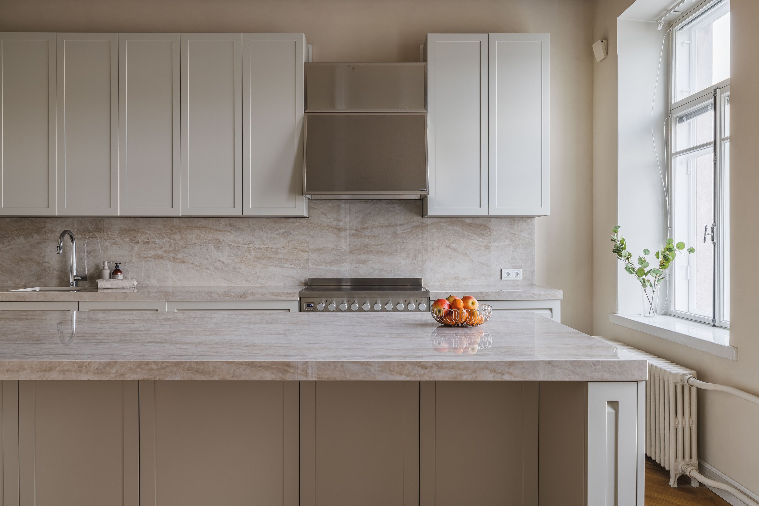 Modern kitchen interior with light gray cabinets, marble countertops, a stainless steel stove, and a bowl of fruit. A large window and a potted plant are on the right.