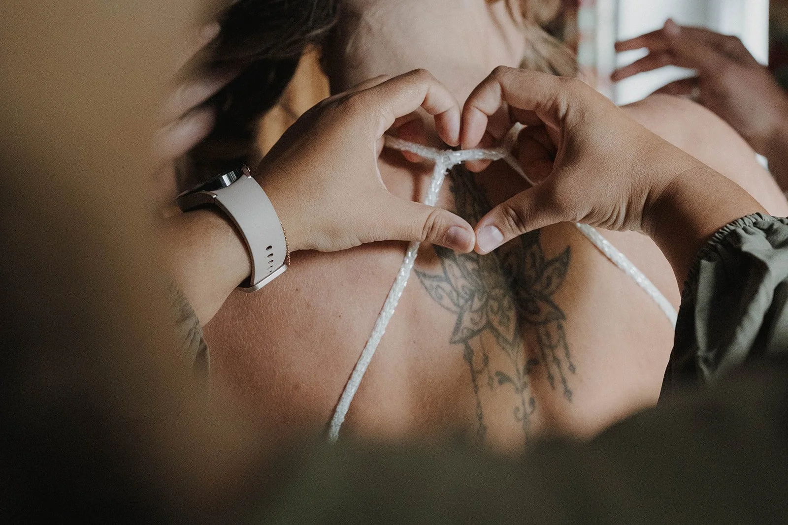 Eine Trauzeugin formt Herz mit den Händen auf der Schulter einer Braut, die ein Tattoo trägt, während diese sich umzuziehen scheint.