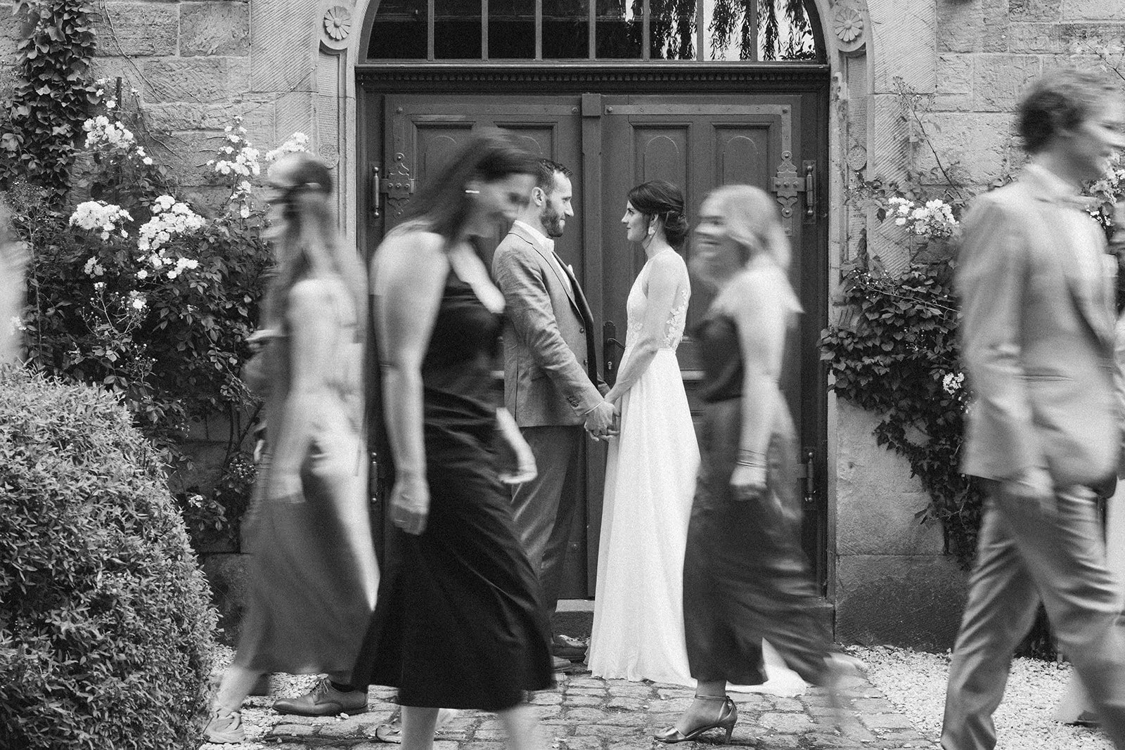 Braut und Bräutigam stehen vor der Tür einer Hochzeitslocation im Rheingau, während Gäste vorbeigehen, unscharfes Bild – festgehalten vom Bart van Westrop, Hochzeitsfotograf in Mainz