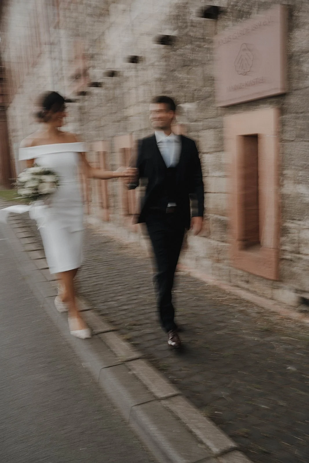 Ein verschwommenes Foto eines Brautpaares, das die Straße entlang geht. Die Braut trägt ein weißes Kleid und hält einen Blumenstrauß, während der Bräutigam einen schwarzen Anzug trägt - festgehalten vom Bart van Westrop, Hochzeitsfotograf in Mainz