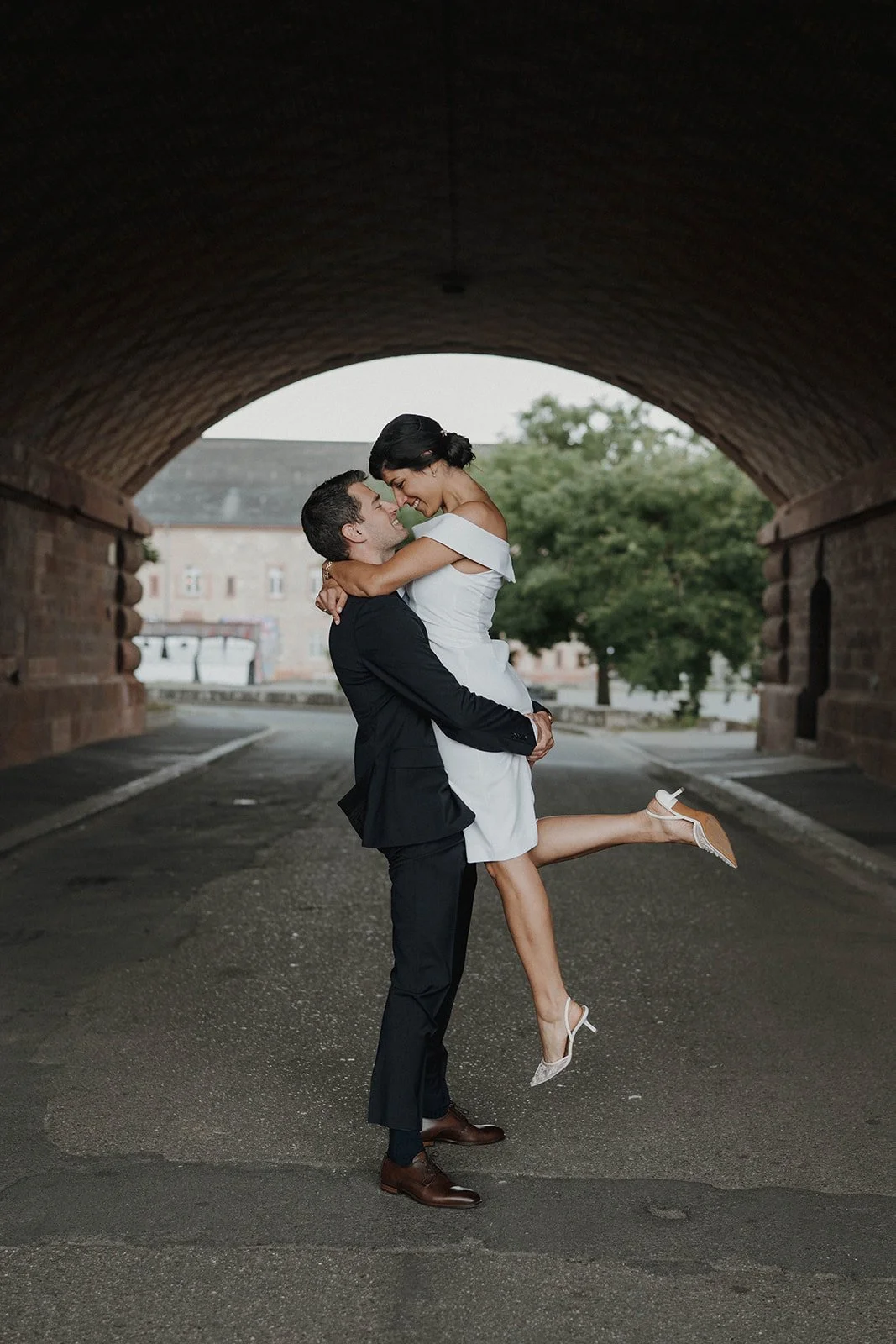 Ein glückliches Brautpaar, das sich küssend auf einer Straße unter einer Brücke befindet. Der Mann hebt die Frau hoch, die ein weißes Kleid trägt - festgehalten vom Bart van Westrop, Hochzeitsfotograf in Mainz