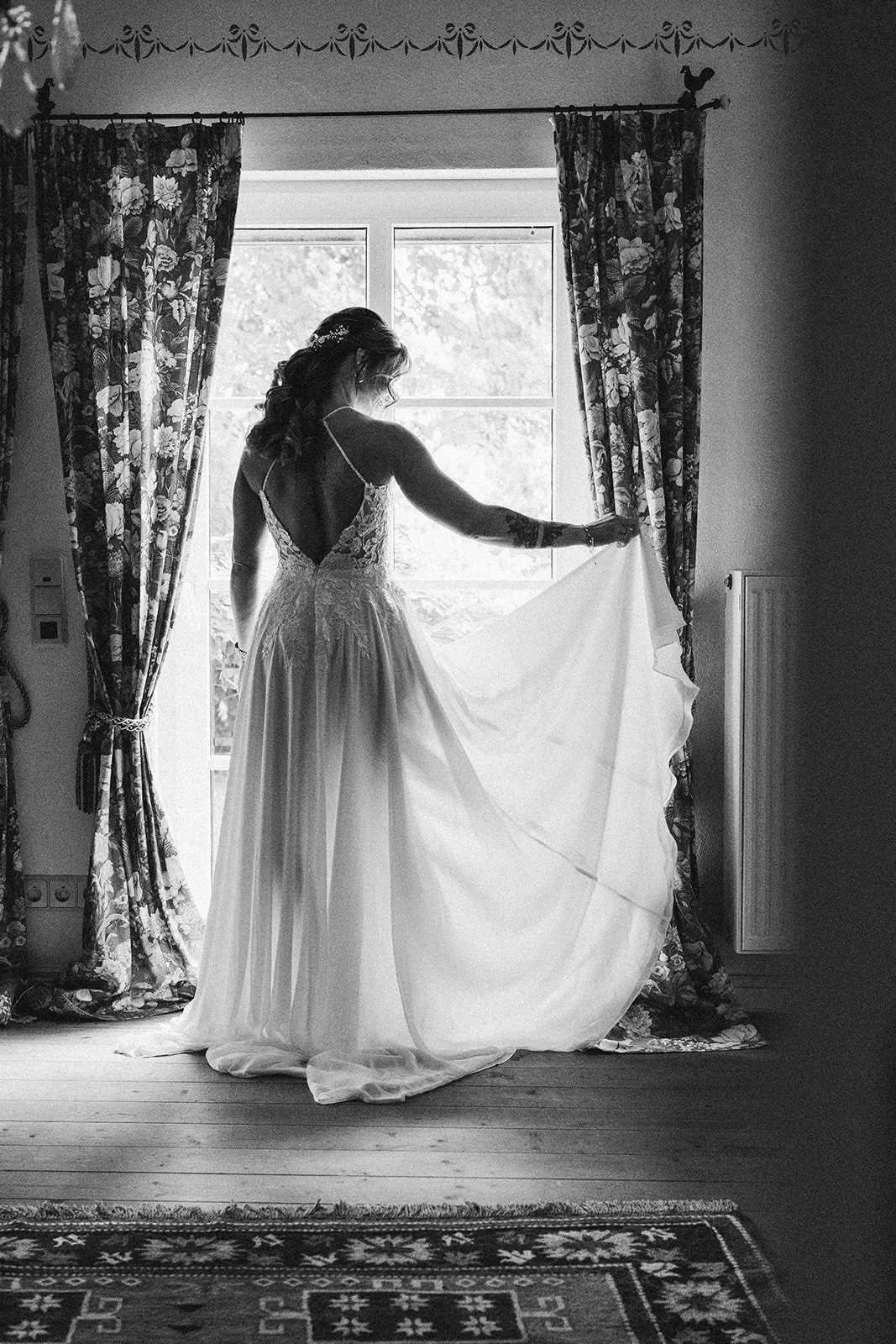 Eine Braut steht vor einem Fenster, hält einen Teil ihres weißen Brautkleides und blickt nach draußen. Das Bild ist in Schwarz-Weiß aufgenommen – festgehalten vom Bart van Westrop, Hoch