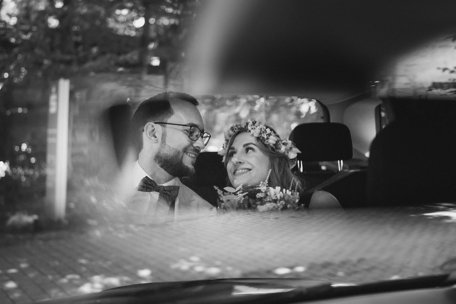 Braut und Bräutigam sitzen im Auto, sehen sich an und lachen, Hochzeit im schwarzen und weißen Stil, mit Blumenkranz und Brautstrauß – festgehalten vom Bart van Westrop, Hochzeitsfotograf in Mainz