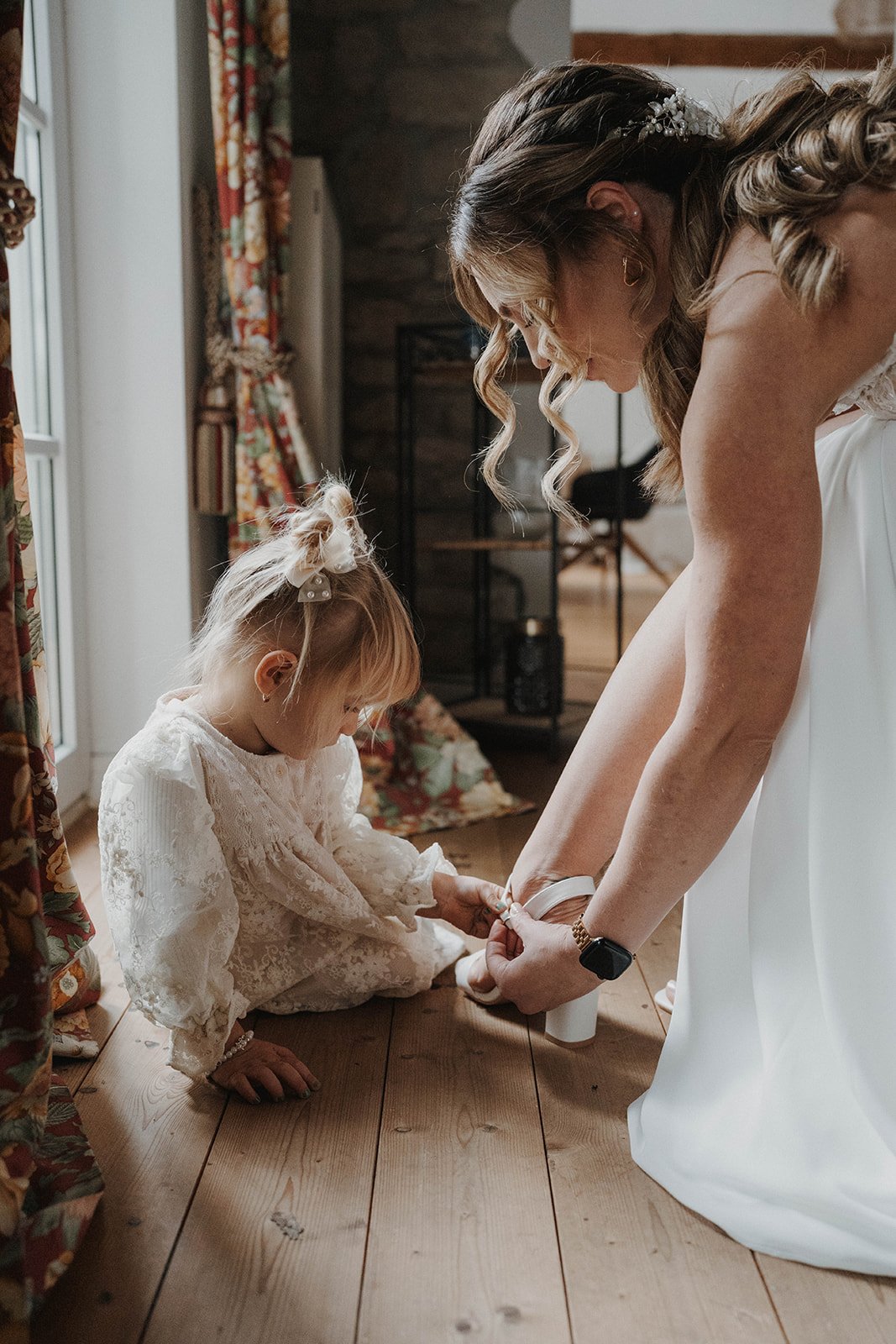 Eine Braut hilft einem kleinen Mädchen, das sich in einem weißen Hochzeitkleid befindet, beim Anziehen ihrer Schuhe in einem Holzraum - festgehalten vom Bart van Westrop, Hochzeitsfotograf in Mainz