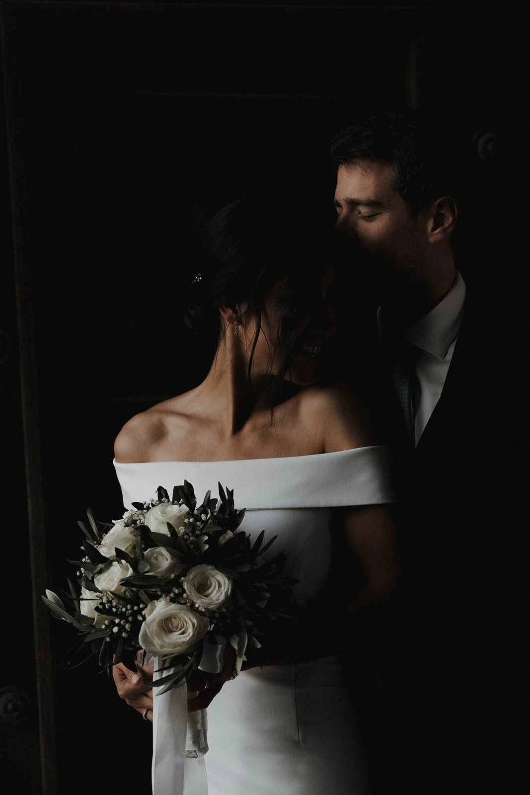 Ein Brautpaar bei einer Hochzeit, die Braut hält einen Blumenstrauß aus weißen Rosen und dunklem Laub, im dunklen Hintergrund – festgehalten vom Bart van Westrop, Hochzeitsfotograf in Mainz