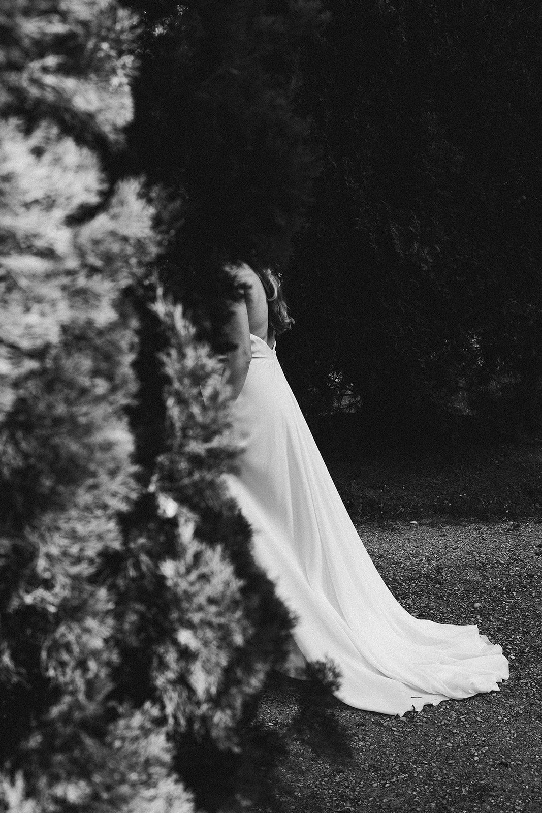 Schwarz-Weiß-Foto einer Frau in weißem Kleid, teilweise hinter einem Baum versteckt, im Hintergrund dunkle Bäume – festgehalten vom Bart van Westrop, Hochzeitsfotograf in Mainz