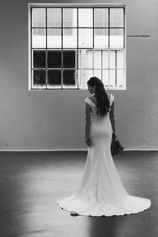 Braut in einem langen weißen Brautkleid steht vor einem großen Fenster in einem Raum mit leeren Wänden, hält einen Blumenstrauß in der Hand und schaut zur Seite.  Hochzeitsfotograf Standesamt Mainz