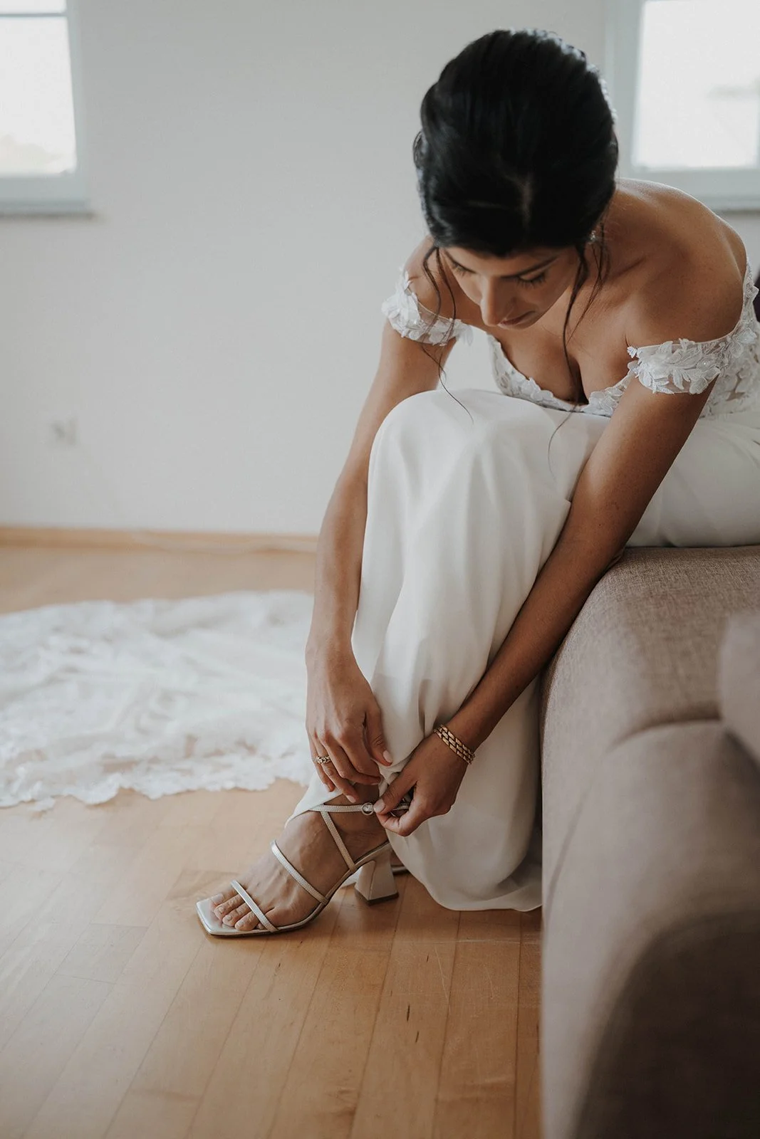 Braut in einem weißen Brautkleid, die ihre Schuhe an einem Holzboden befestigt, während sie auf einer Couch sitzt – festgehalten vom Bart van Westrop, Hochzeitsfotograf in Mainz
