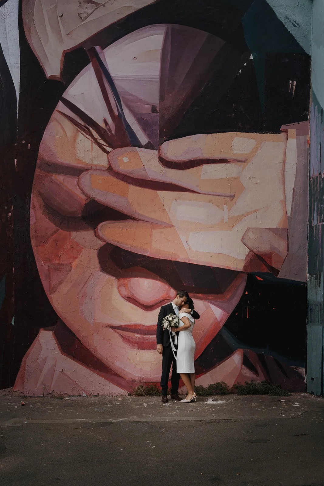 Ein verliebtes Paar in formeller Kleidung, die sich umarmen, vor einer großen Wand mit Street Art. Die Wand zeigt ein abstraktes Porträt eines Gesichts, das mit geometrischen Formen und warmen Farbtönen gestaltet ist.