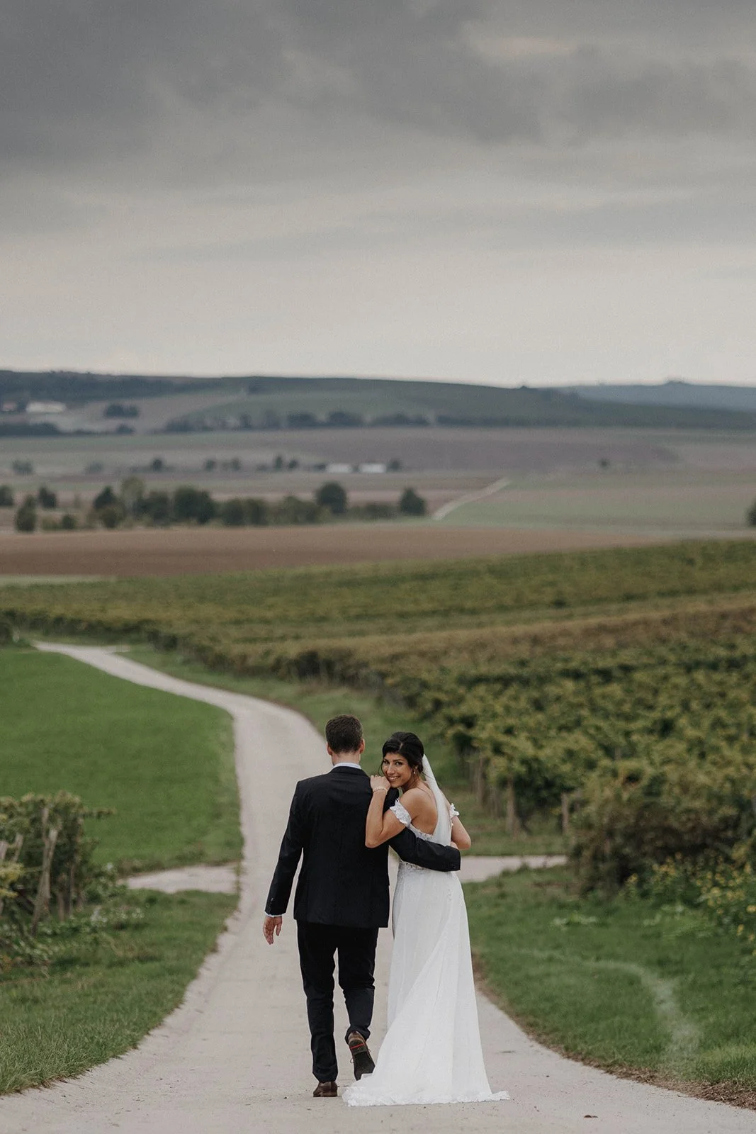 Ein Brautpaar läuft Hand in Hand auf einem Landweg in einer landwirtschaftlichen Landschaft. Die Braut trägt ein weißes Hochzeitskleid und der Bräutigam einen schwarzen Anzug.- festgehalten vom Bart van Westrop, Hochzeitsfotograf in Mainz