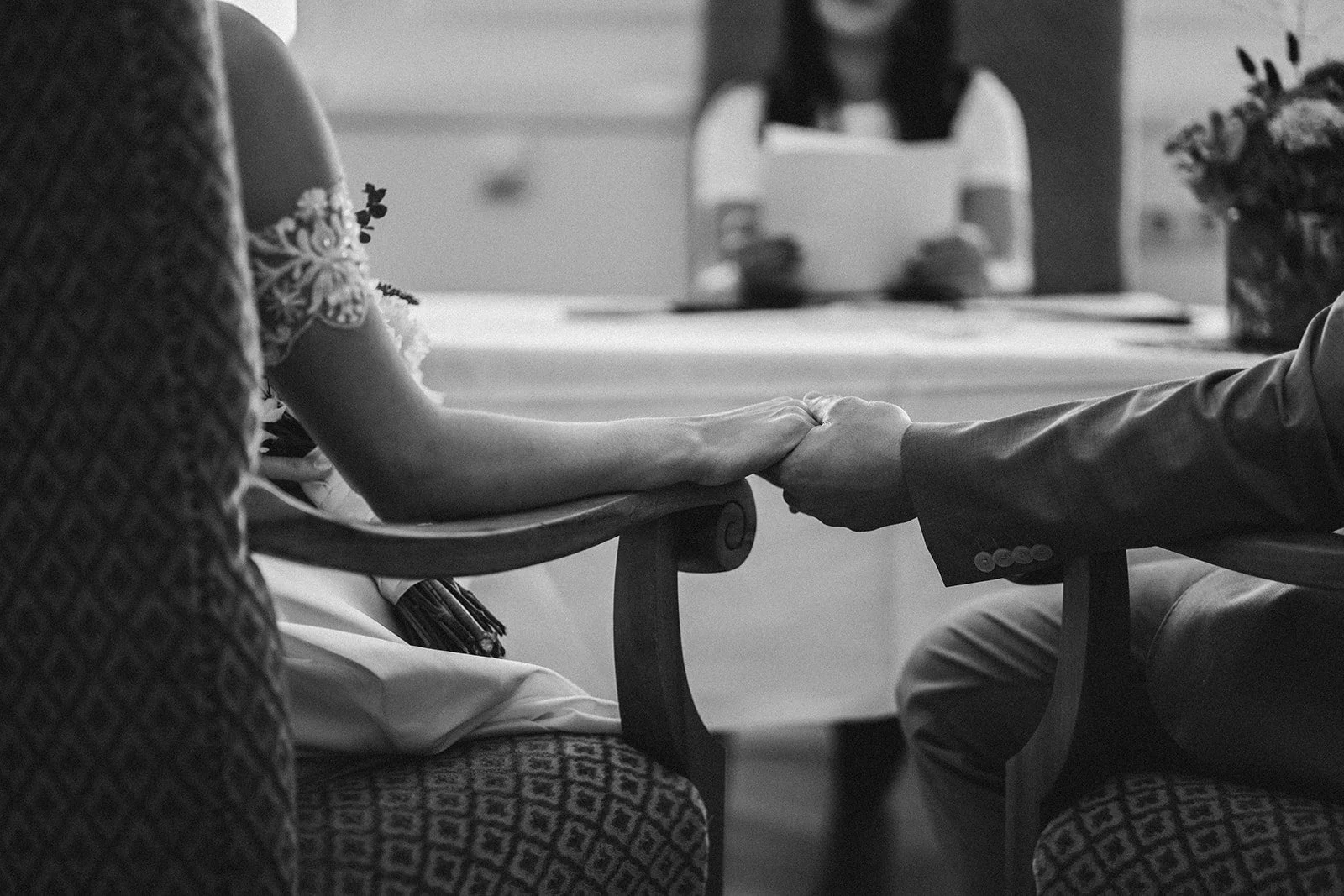 Schwarz-Weiß-Foto von einem Paar, das bei einer Hochzeit die Hände hält, während sie auf Stühlen sitzen, im Hintergrund ein Kind in einem Hochstuhl.  Hochzeitsfotograf Standesamt Mainz