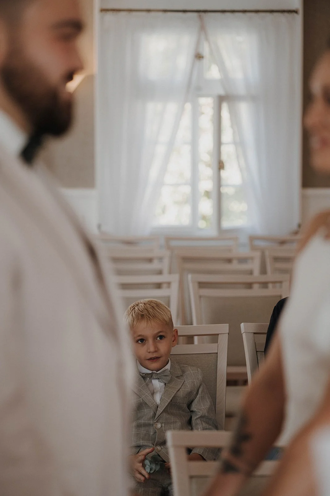 Ein kleiner Junge in einem grauen Anzug sitzt in einem Raum mit vielen Stühlen. Hochzeitsfotograf Standesamt Mainz