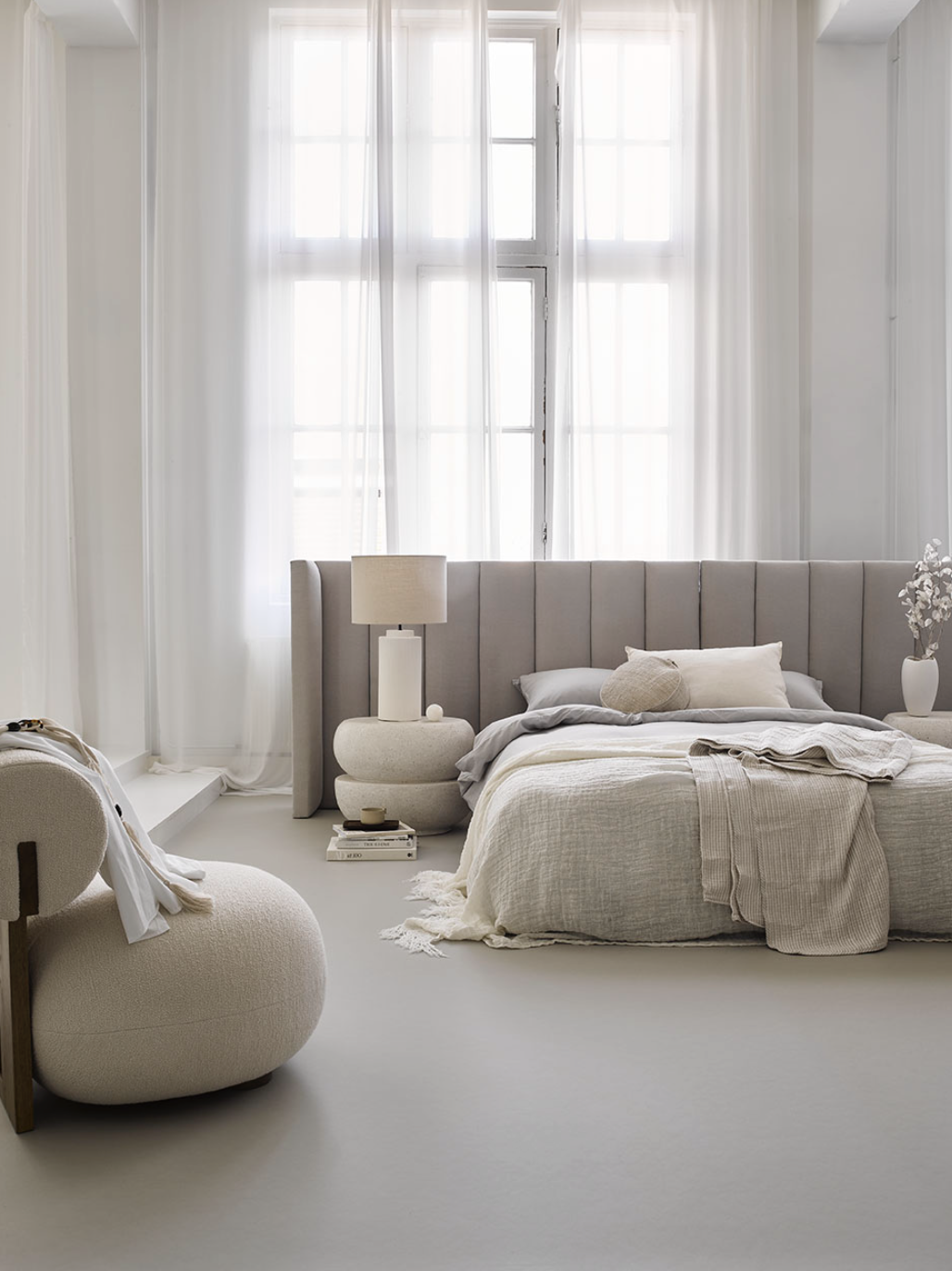 Een minimalistische slaapkamer met witte wanden, grote ramen met witte gordijnen, een bed met beige bekleding en beddengoed, een bijzettafel met een lamp en boeken, en decoratieve vazen met bloemen.