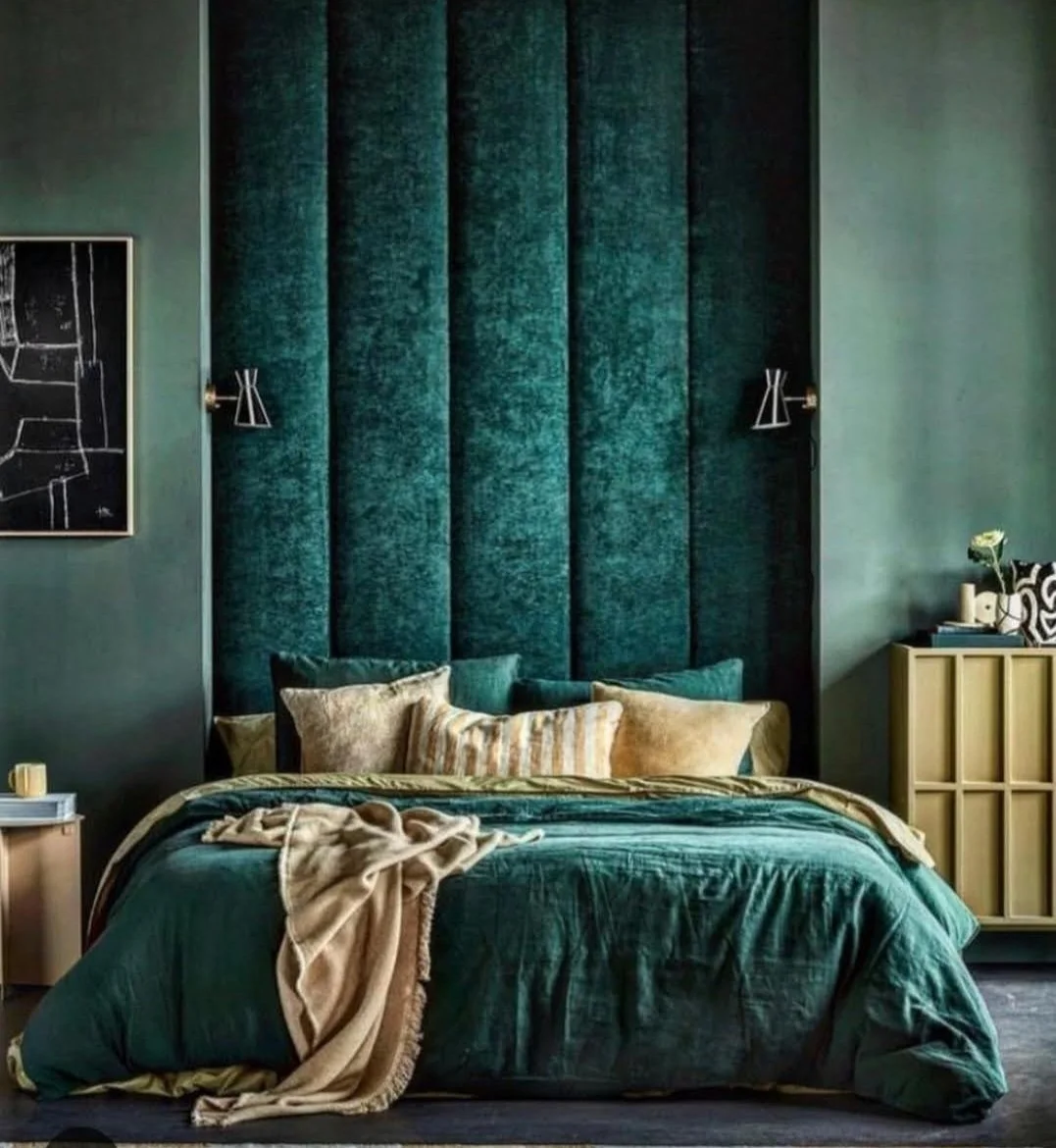 Een slaapkamer met een donkergroene, padded hoofdwand, een bed met groene beddengoed en verschillende kussens, een beige plaid en een neplugge kast met decoratieve items, een muurkunstwerk en wandlampen.