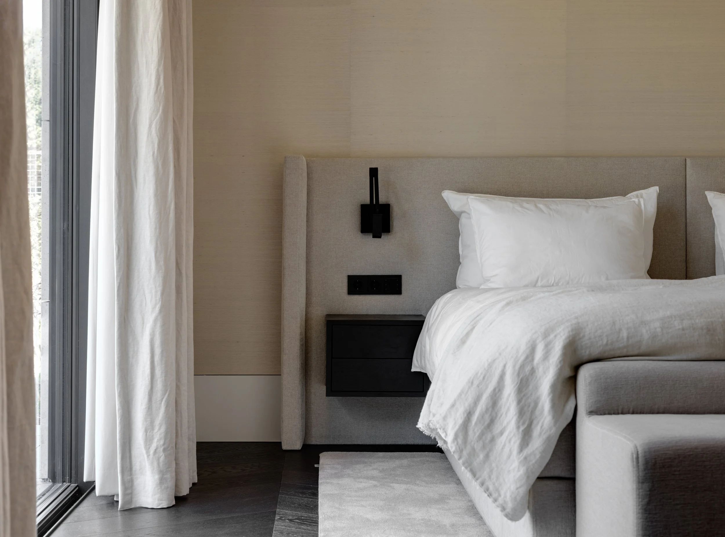 Een slaapkamer met een groot bed, witte lakens en kussens, een beige hoofdbord en een zwart nachtkastje met een lamp en stopcontacten.